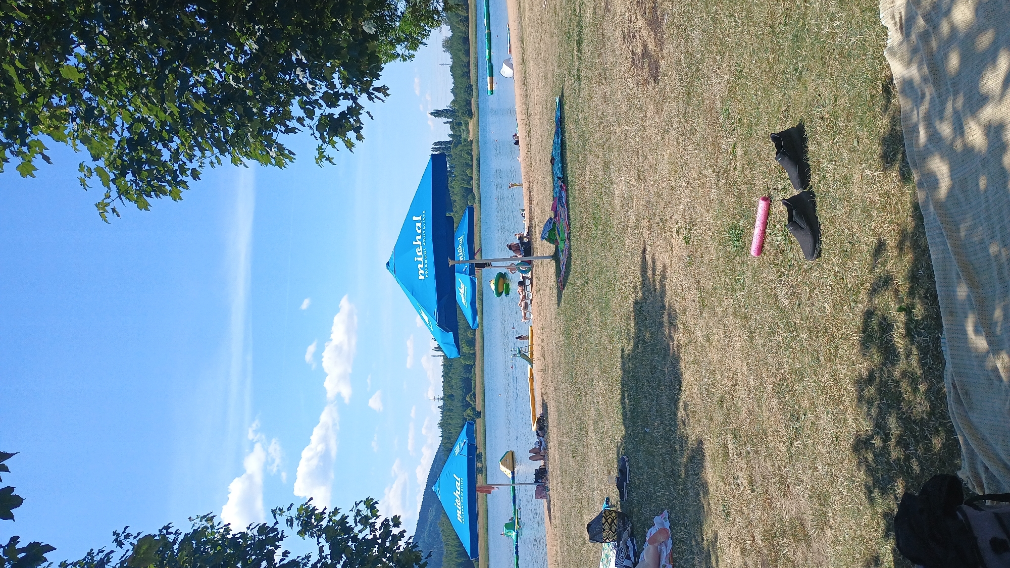 The image depicts a sunny day at Michal's Pond (Michalský rybník) in Sokolov, Czechia. The scene captures a lakeside recreational area with a sandy shore and grassy embankment. Several blue beach umbrellas with the "michal" logo are set up. People are visible on the beach and in the shallow water, some lounging on towels and others near inflatable water attractions. In the foreground, on the dry grass, a pink pool noodle and a pair of black slip-on shoes are placed. Trees frame the top of the image, with branches and leaves casting shadows on the ground. The lake stretches into the background, bordered by a line of trees and a distant hill. The sky is bright blue with scattered white clouds.