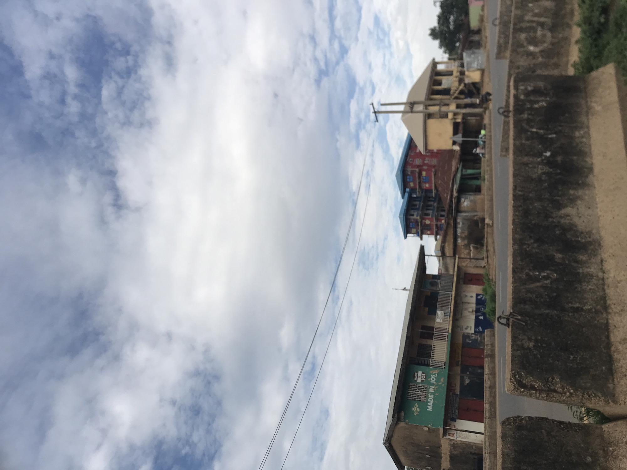 The image displays an urban street scene in Jos, Nigeria, under a predominantly cloudy sky with patches of blue. In the immediate foreground, a line of weathered concrete road barriers, dark grey with some visible grime and faint graffiti, separates the viewer from the roadway. A gap between two barriers on the left reveals some overgrown vegetation.
Beyond the barriers lies a paved asphalt road, which appears to be largely clear of active traffic. On the far side of the road, several buildings line the street.
To the left, a two-story building with a corrugated metal roof is prominent. Its upper floor is light brown/tan, while the lower section features a teal/green facade. A white sign on the green section explicitly reads "MADE IN JOS," with smaller text above it including "MADE IN JOS" again and "SALE," alongside a small graphic. The upper floor has a white-railed balcony and windows with metal grates. The ground-floor storefronts are closed, characterized by red and blue metal shutters. Overhead, multiple black power lines traverse the sky from left to right.
Further to the right and slightly behind, a cluster of multi-story buildings, possibly under construction or recently completed, is visible. One distinct structure features red walls with blue window frames and a blue sloped roof. Another similar blue-roofed building is partially obscured behind it.
To the far right, another two-story building stands with a light yellow/tan exterior and a light brown/tan pitched roof. An external staircase, possibly wooden, is visible on its left side, leading to the upper floor. A wooden utility pole, supporting power lines, is positioned in front of this building. In the mid-ground, near the yellow building, two small vehicles, likely motorbikes or mopeds, are visible on the roadside, and a faint human figure can be discerned nearby. Sparse green vegetation is present along the roadside and a dark-foliaged tree is visible on the far right edge of the frame.