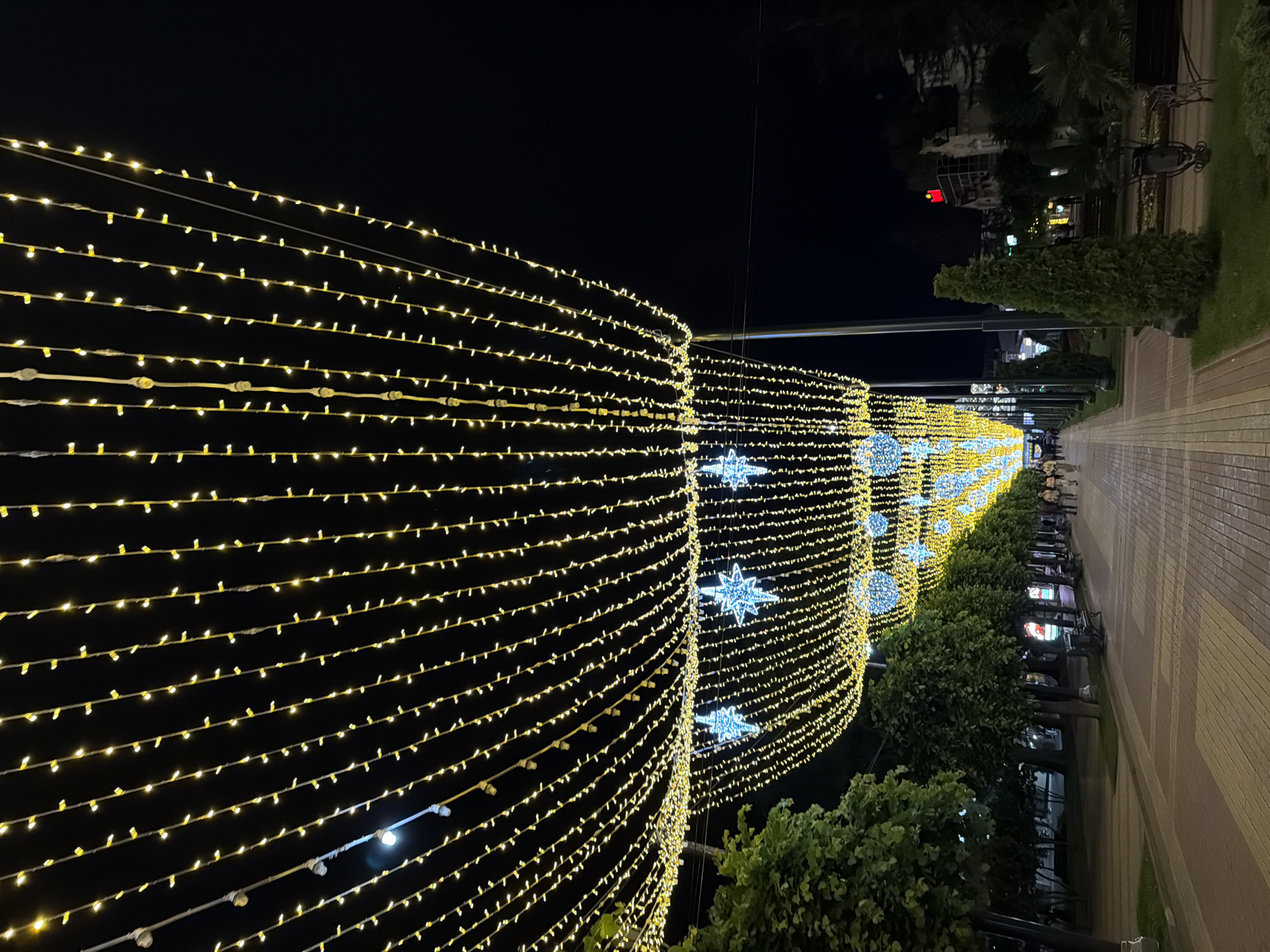 The image shows a pedestrian street at night in Kislovodsk, Russia, illuminated by strings of yellow lights and large star-shaped light decorations. The street is paved with light-colored tiles and bordered by trees on the left and benches and greenery on the right. A few people are visible walking on the street. The overall scene suggests a festive or celebratory atmosphere.