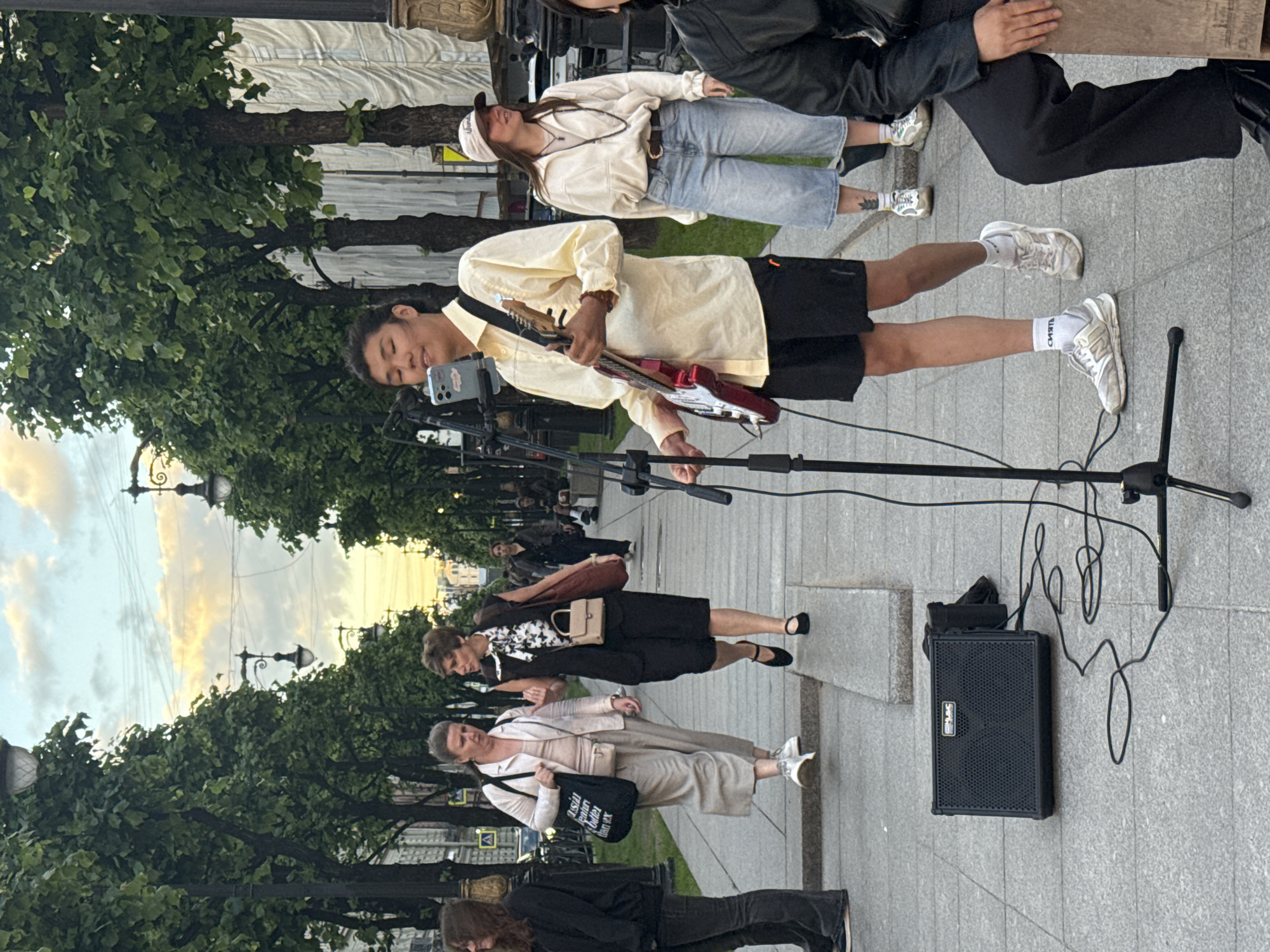 An Asian male street performer plays a red electric guitar and sings into a microphone on a paved walkway lined with trees. He is wearing a yellow button-down shirt, black shorts, and white sneakers. A small black amplifier is on the ground in front of him, with cables connecting it to the microphone stand. Behind him, two women walk past, one in a beige jacket and skirt, the other in a dark vest and shorts. A young woman wearing a beige jacket and denim shorts stands to the right of the performer. The scene is set in Saint Petersburg, Russia, as indicated by the architecture and streetscape visible in the background. The sky is cloudy with hints of sunset colors.