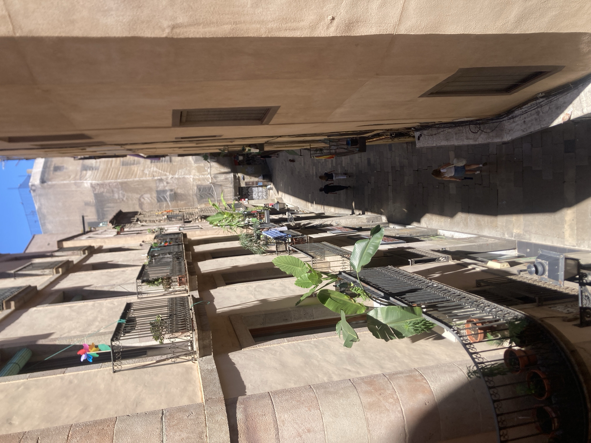 An eye-level long shot, vertically oriented, depicts an urban alleyway between tall buildings, potentially in Barcelona, Spain, based on architectural style. The alleyway is paved with grey stone blocks. Two people can be seen walking on the right side of the image, a woman wearing shorts walking away from the camera. A man walking perpendicular to the camera. The buildings are cream-colored with closed, shuttered windows and wrought-iron balconies. Plants are visible on several of the balconies. The architecture is old-style, with stone and stucco surfaces. A colorful pinwheel is seen attached to a balcony. The light suggests a sunny day, with some areas in shadows. The alleyway recedes into the distance.