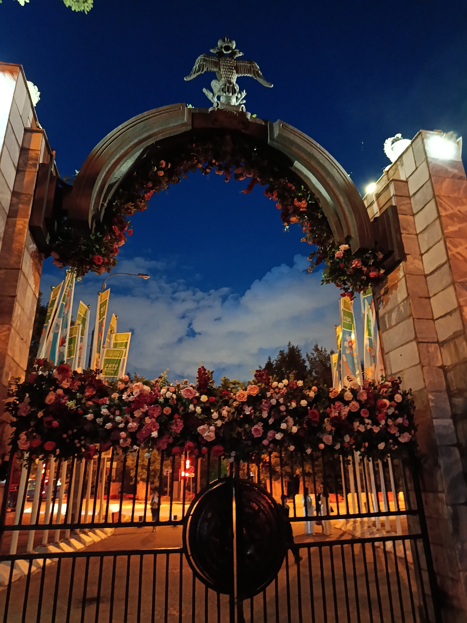 The image depicts a grand, ornate archway at night, likely serving as an entrance in Moscow, Russia. The archway features robust stone pillars supporting a large, curved metal or stone arch. A prominent statue of an eagle with outstretched wings, possibly a double-headed eagle, crowns the top of the arch.
The structure is heavily decorated with an extensive display of artificial flowers, primarily roses, in various shades of pink, red, and white, with accompanying green foliage. These floral garlands adorn the top and sides of the arch, cascading down.
Through the archway, several tall, rectangular banners or flags are visible. On one banner, the Cyrillic text "АЭТО КЛУБ" (Aero Club) is clearly legible, suggesting the identity of the establishment or event. The background reveals a deep blue night sky dotted with scattered white clouds.
At the bottom, a black metal gate with vertical bars is partially open, offering a glimpse of the scene beyond. Blurry figures of people and vehicles are visible on a street or pathway, illuminated by the warm orange glow of distant streetlights and traffic lights. The overall ambiance is festive and grand, captured under the artificial illumination of an urban evening.