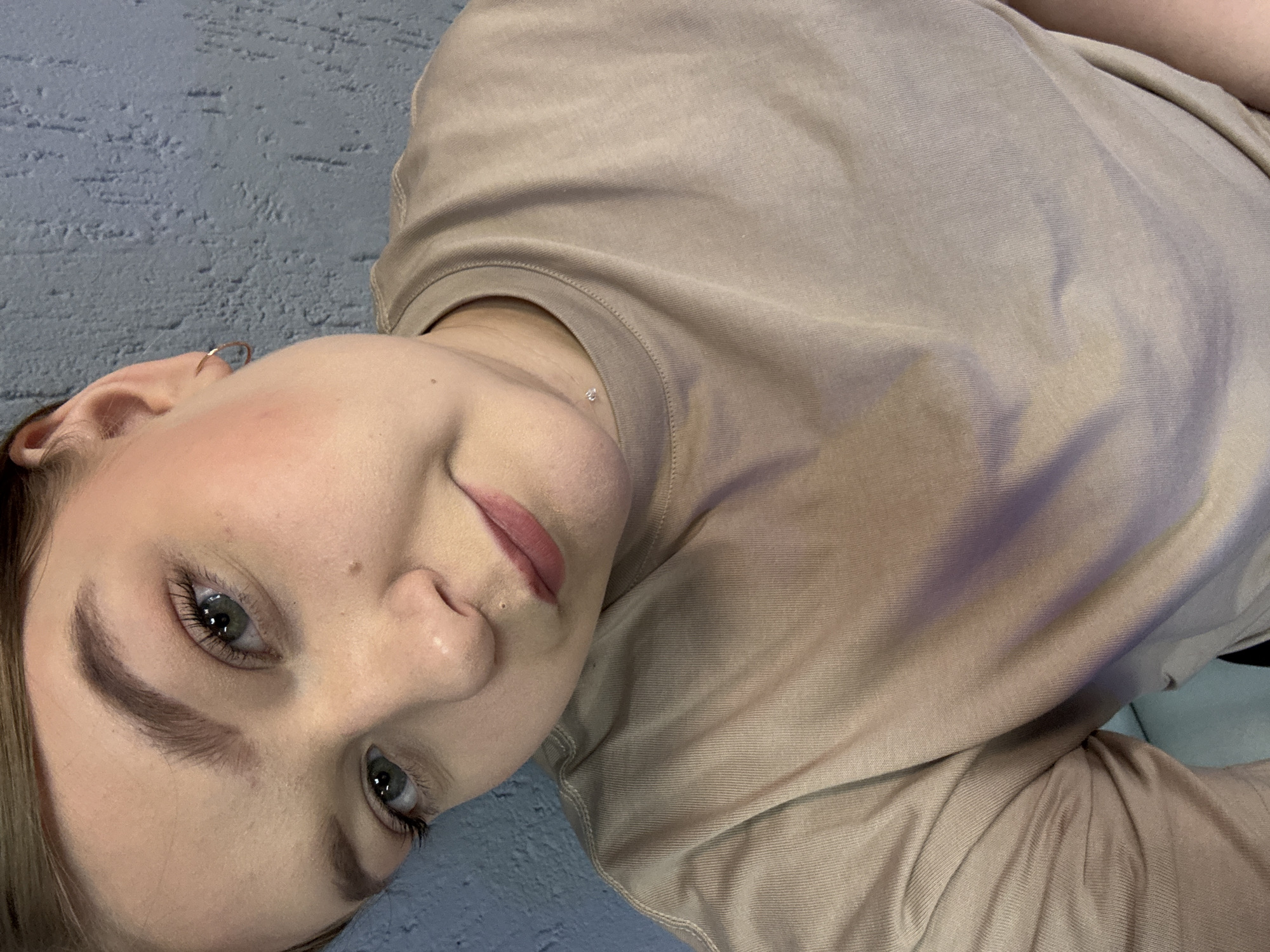 An indoor close-up photograph depicts a young adult female, appearing to be Caucasian, with light skin, light brown hair, and green eyes, looking directly into the camera with a subtle smile. Her head is tilted slightly. She has groomed brown eyebrows, subtle pink blush on her cheeks, and pink-tinted lips. A small, metallic stud piercing is visible on the right side of her nose, superior to the nostril. She wears a plain, light beige crew-neck t-shirt. A small, gold-toned hoop earring is present in her visible right earlobe, and a small, clear, faceted stud pendant is suspended from a thin chain around her neck. The background consists of a textured, matte, blue-grey wall, suggestive of a plaster or stucco finish. The lighting is soft and even. The specific location within Chaikovsky, Russia, is not ascertainable from the visual content of the image.