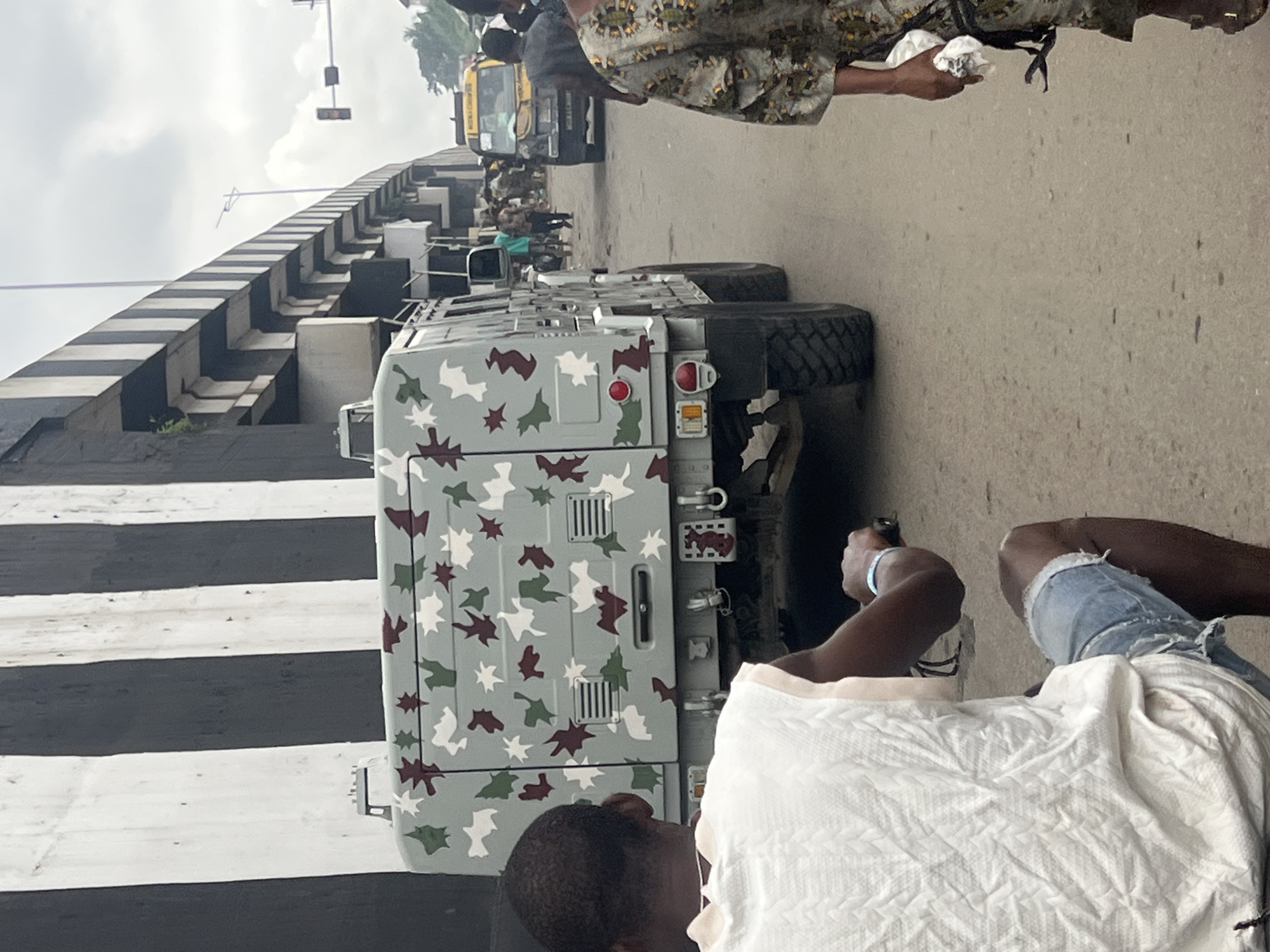 In Nkpor, Nigeria, under a cloudy sky, a military-style vehicle with camouflage paint is parked on a road. The vehicle is large, with thick tires and appears to be an armored personnel carrier. The camouflage pattern consists of irregular shapes in white, green, and burgundy. Behind the military vehicle, a yellow taxi is visible, along with other vehicles and people walking in the background, suggesting a busy street scene. An overpass with black and white striped pillars looms above the road, casting shadows. In the foreground, a person wearing a white sleeveless top and ripped denim shorts is partially visible from behind, seated on what appears to be a motorcycle or similar vehicle. To the right, another person in a camouflage outfit is walking away from the camera. The overall impression is a street in a Nigerian city, with a mix of civilian and military presence.
