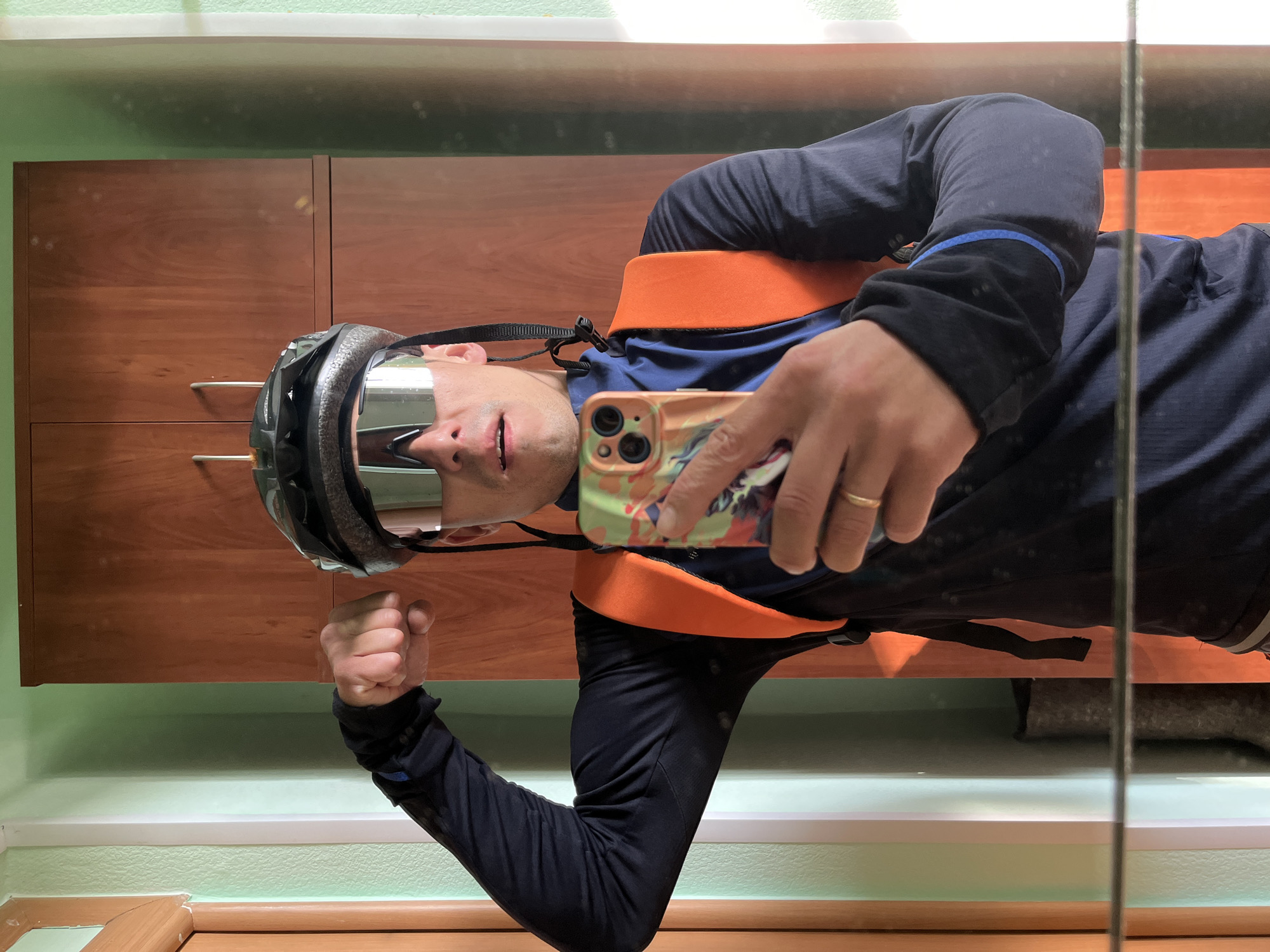 A man is taking a selfie in front of a mirror. He is wearing a cycling helmet and mirrored sunglasses. He has on a dark blue long-sleeved shirt with a blue stripe on the sleeve and an orange sleeveless vest. He is holding a phone in his right hand, which is visible in the mirror, and has a wedding ring on his finger. His left fist is clenched. Behind him, the room has brown cabinets and a pale green wall with white trim. The location is difficult to ascertain, but given the city reference, it is likely an interior space in Chaikovsky, Russia.