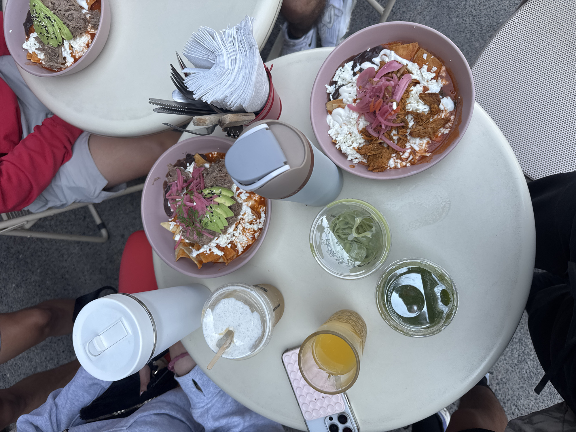 The photograph depicts a table setting with various items on a white round table. There are three bowls of what appear to be loaded tortillas with toppings like meat, cheese, avocado, and pickled onions. Next to the bowls are a red holder with cutlery and napkins, several beverages in cups and tumblers, and a mobile phone. People are seated around the table, as evidenced by the portions of clothing and legs visible in the frame. The scene appears to be outdoors, possibly at a restaurant or cafe. Based on the food and the assumption it is a Puebla, Mexico restaurant, this image most likely depicts Chilaquiles Poblanos or a variant of it.