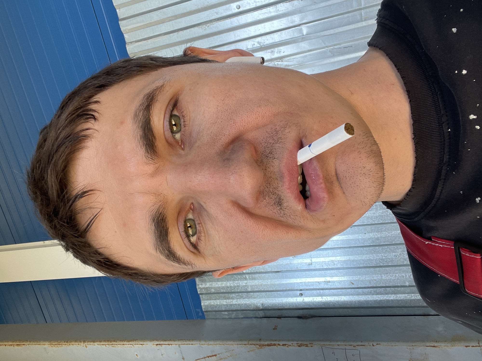 A man with short, dark hair, green eyes, and light stubble on his face is looking directly at the camera. He is holding a cigarette in his mouth. He is wearing a black t-shirt with white speckles and red suspenders. He has a white earbud in his right ear. The background consists of corrugated metal siding and a blue striped structure. The scene appears to be outdoors, possibly in an industrial or utilitarian area.