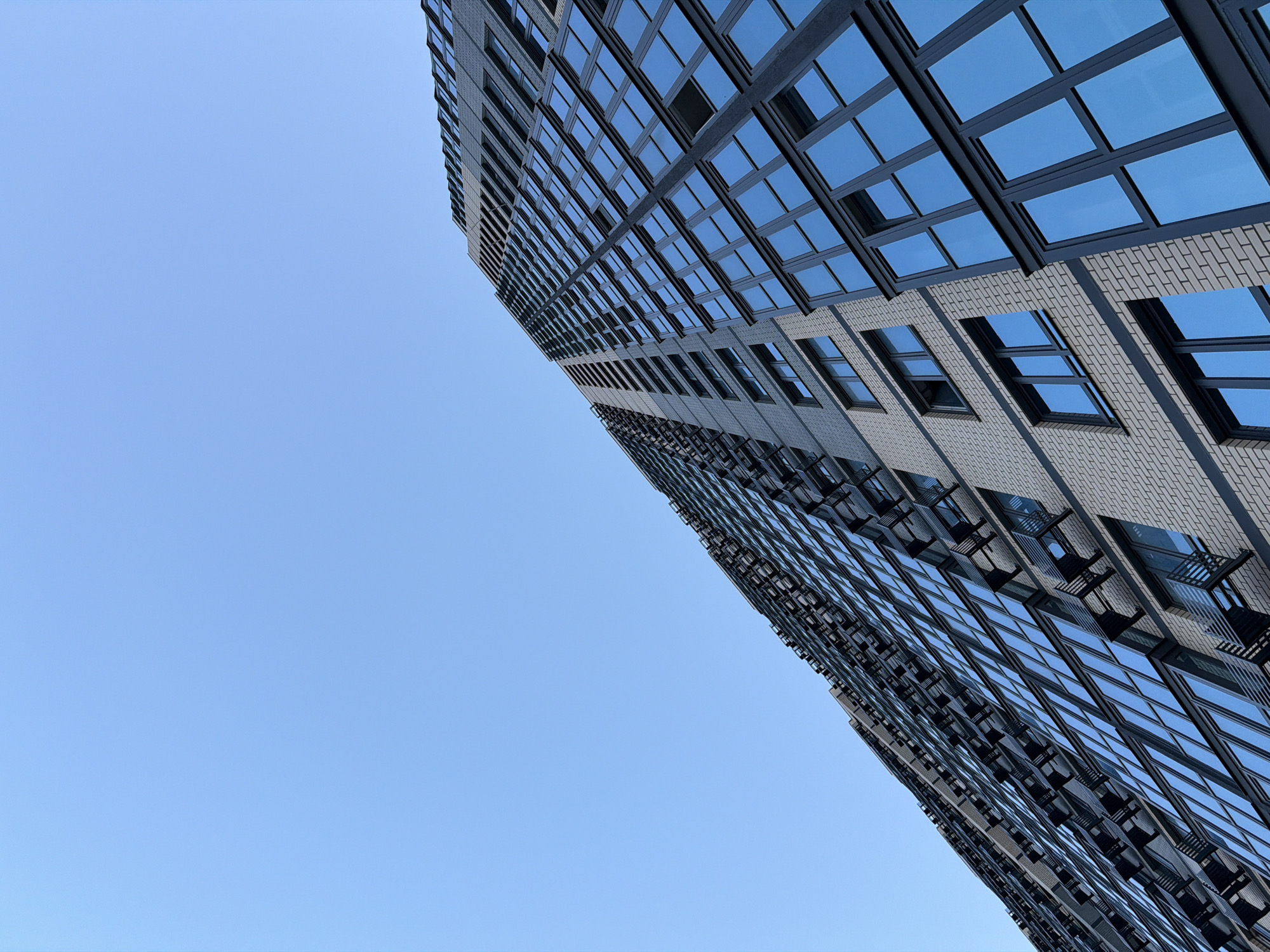 A modern, multi-story building dominates the right side of the frame, captured from a low-angle perspective looking upwards. The structure is characterized by a facade that appears to alternate between light grey or white brickwork and expansive sections of dark-framed windows. The windows are numerous, reflecting a clear, bright blue sky, suggesting daylight. The building extends diagonally from the lower right to the upper left corner, creating a dynamic composition that emphasizes its height.
The sky occupies the majority of the left and upper-middle portions of the image, presenting a uniform, cloudless expanse of light blue. The reflections in the windows are consistent with this clear sky. Some sections of the building appear to have small, dark protrusions or awnings above certain windows, particularly in the lower visible areas. The architecture suggests a contemporary design, likely a residential or commercial high-rise in an urban setting. No specific activity or individuals are visible. The image captures a serene, bright day in what is contextually identified as Beryozovy, Russia. There is no visible text.