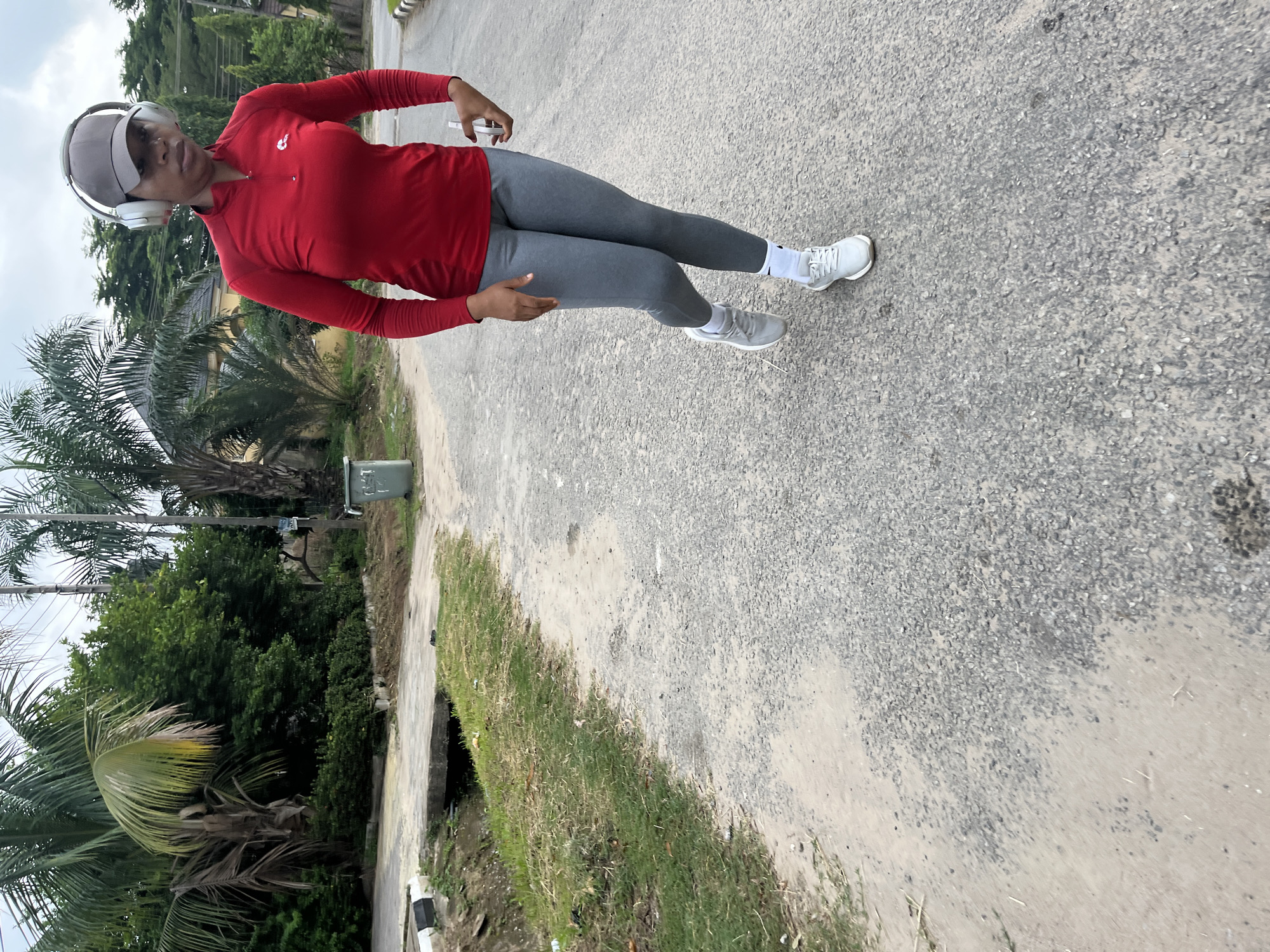 An outdoor, full-body vertical shot features a person in athletic wear on a paved surface, set against a backdrop of lush greenery. The individual, appearing to be female, stands facing slightly to the viewer's right, wearing a red long-sleeved athletic top with a small white logo on the chest, grey full-length leggings, white socks, and white athletic shoes. A grey baseball cap covers her head, and white over-ear headphones are worn over the cap. Her left arm is slightly bent, and her right hand holds a white rectangular object, possibly a smartphone.
The ground beneath her feet is a mix of dark grey asphalt or tarmac and lighter, possibly sandy or dusty patches. To the left, a narrow strip of green grass borders the paved area, with a concrete curb and what appears to be a drainage ditch. In the background, numerous vibrant green trees and several distinct palm trees indicate a tropical or subtropical climate, consistent with Abuja, Nigeria. A light-colored building is partially visible behind the trees, and a grey utility box or small bin is situated near the left edge of the frame. The sky is overcast, suggesting daytime with subdued lighting. The person appears to be pausing during a walk or exercise session, exhibiting a neutral, focused expression. No distinct readable text is visible on clothing or objects, other than a vague logo on the top.