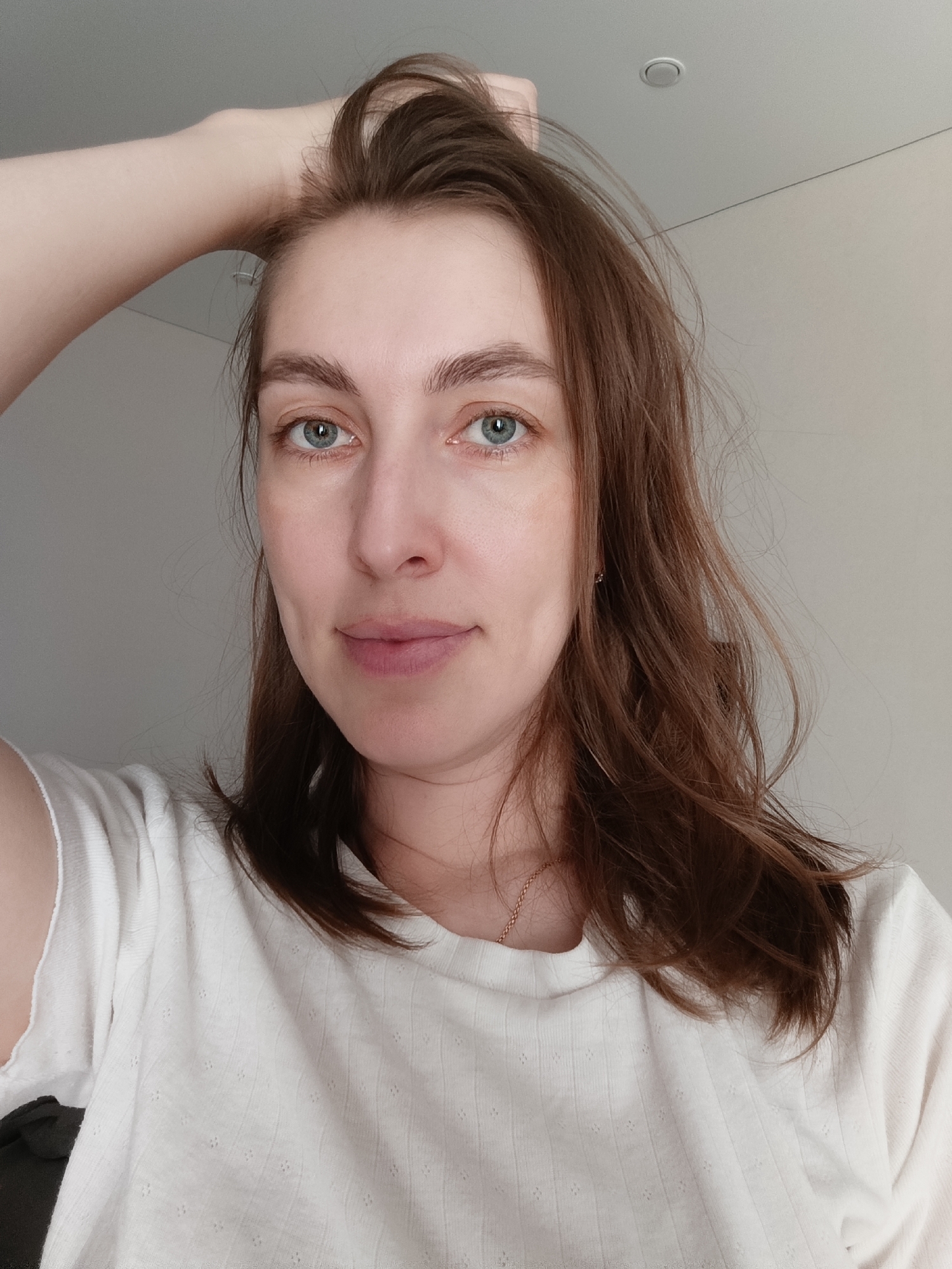 A woman with brown hair and blue eyes is taking a selfie indoors. She is wearing a white t-shirt. Her left hand is running through her hair. The background is a plain white wall with a recessed lighting fixture. The location is Chaikovsky, Russia.