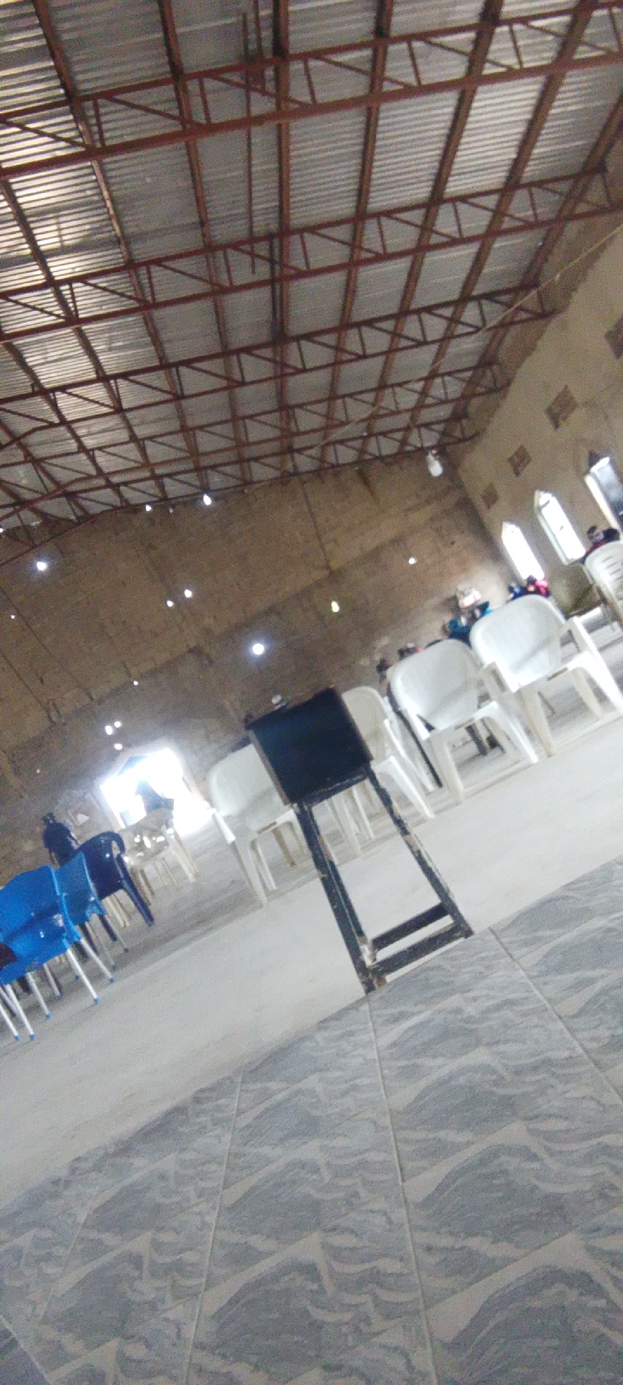 The image shows the interior of a large, sparsely furnished room with a corrugated metal roof supported by a red metal truss system. The walls are made of rough, light-colored material. The floor is tiled with a gray and white patterned tile.
In the foreground, there are several white plastic chairs arranged in rows, and a black object that appears to be a stand or small table with a black top. Further back, there are more white plastic chairs, some blue plastic chairs, and a few people are visible. One person is standing near a bright light source in the background, which appears to be an entrance or opening. Another person is seated in a white chair in the middle ground. To the right, there are windows with arched tops.
The overall impression is that of a community hall or a simple place of worship that is either being set up or has recently been used. The lighting is uneven, with bright spots near the entrance and general illumination from overhead lights. The perspective is from a low angle, looking upwards towards the roof and across the room.