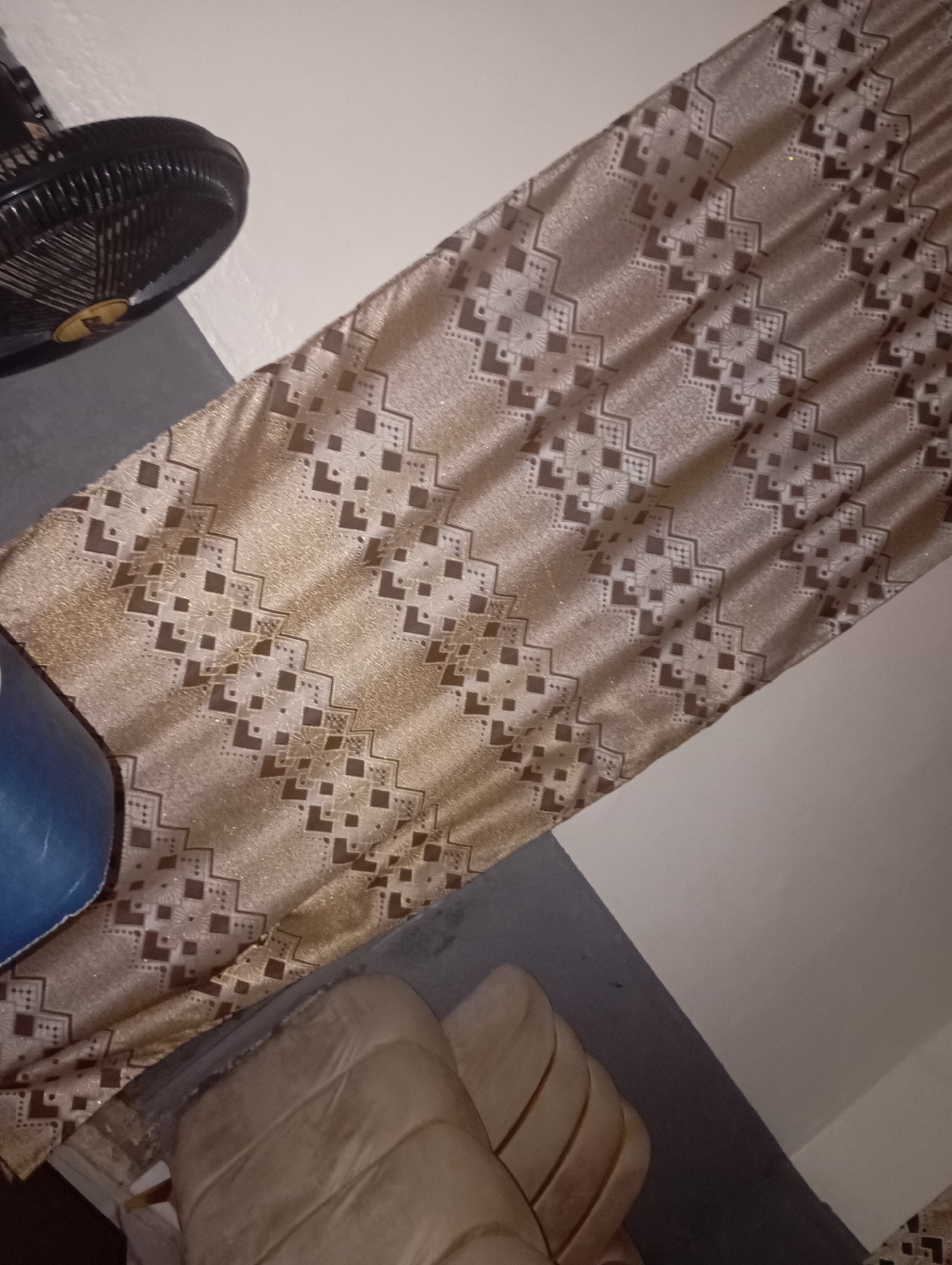 The image depicts a close-up, slightly tilted view of a room. In the upper left corner, a black oscillating fan is partially visible. Dominating the center of the frame is a length of fabric with a gold-toned shimmering base and a repeating geometric pattern in brown. The fabric appears to be draped or hung, creating folds and shadows. In the lower portion of the image, a portion of a light-colored, plush object, possibly a sofa or chair, is visible. The background consists of a wall with a distinct color change, suggesting two different painted sections or a corner. The lighting is dim, with some areas appearing brighter due to the fabric's sheen. The scene is indoors and does not contain any discernible elements that would specifically place it in New Stadium, Nigeria.