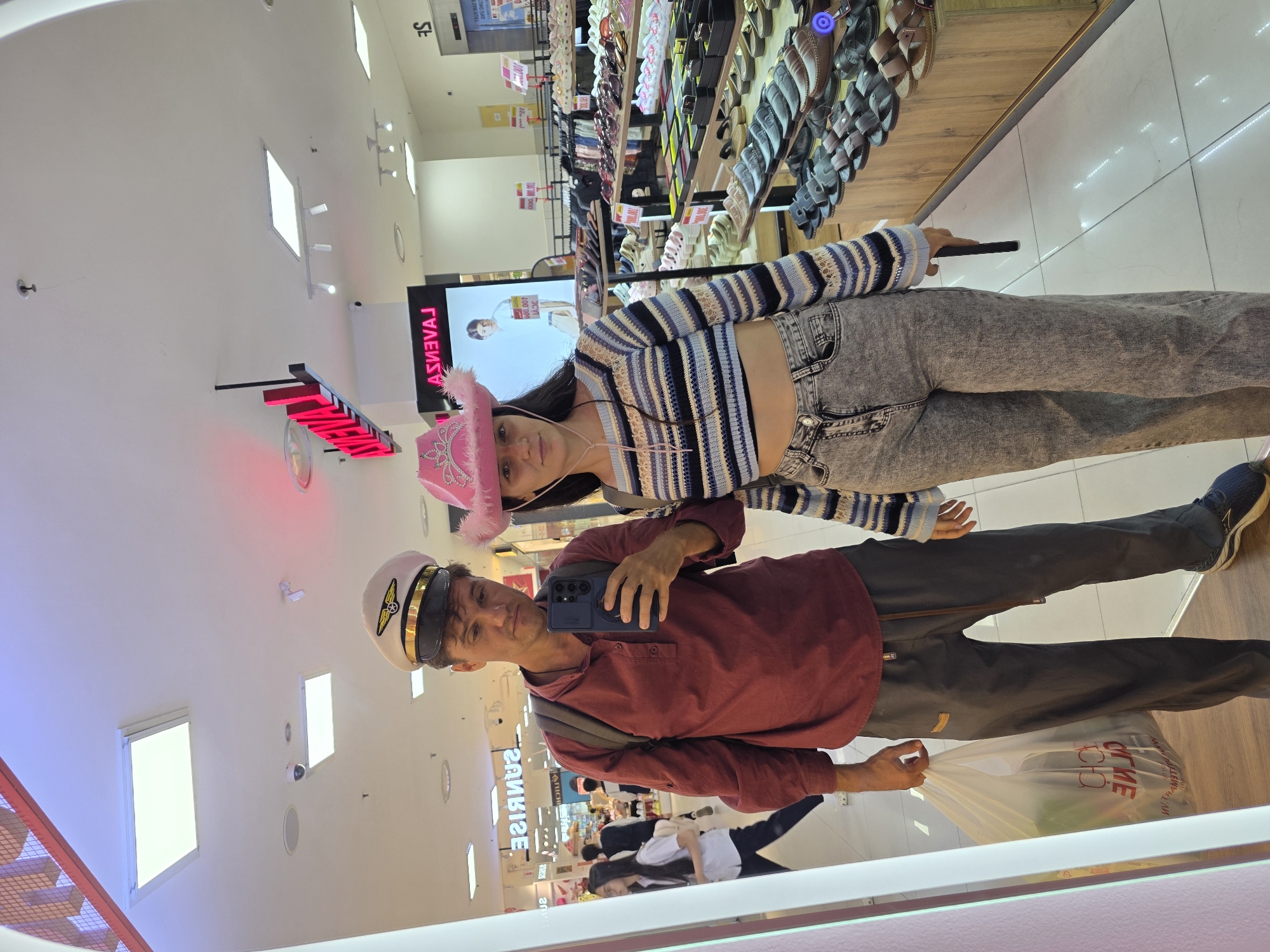 The image shows a mirror reflection of two people posing in what appears to be a shopping mall. The man is wearing a captain's hat, a red shirt, and dark pants, holding a phone in one hand and a shopping bag in the other. The woman is wearing a pink cowboy hat, a blue and white striped top, and gray jeans. Behind them, shelves filled with shoes are visible. The reflection also shows store signs and people walking in the background. Signs indicating "LEWE," "CO," and "ASEA," as well as price tags with amounts such as "2.000" and "600.00," can be seen. The location is likely in Phú Thủy, Vietnam, based on the store signage and visual cues.