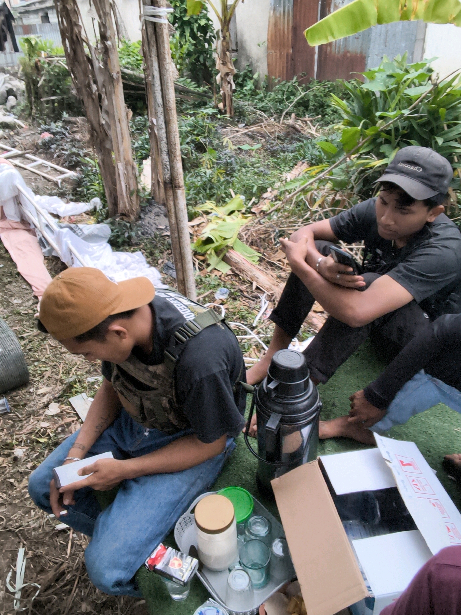In an outdoor urban setting, possibly within the city of Sungai Penuh, Indonesia, two young men are seated on the ground amidst debris and vegetation. The ground is covered with a green matting in the foreground, and a mix of dirt, leaves, and discarded items in the background. Several banana plants are visible, along with tin-paneled structures, suggesting a mixed rural and urban environment.
One man, wearing a black t-shirt, black pants, and a black baseball cap, is seated on the green matting, holding a smartphone. He is looking down at the device. The other man, wearing a brown baseball cap, a black t-shirt, a tactical vest over his t-shirt, and blue jeans, is crouched down, holding a white object, possibly a packet or a small box. His attention is focused on the object in his hands.
In the foreground, a large black thermos sits on the green matting, next to a glass table. On the table are several glasses, a jar with a beige lid containing a white substance, and a pack of cigarettes. A cardboard box is also present on the table. The overall scene appears informal and candid, depicting a moment of rest or engagement with personal items in an outdoor, somewhat unkempt, environment. The presence of the tactical vest on one individual might indicate a role in security or a specific activity, but without further context, it remains a speculative detail.
