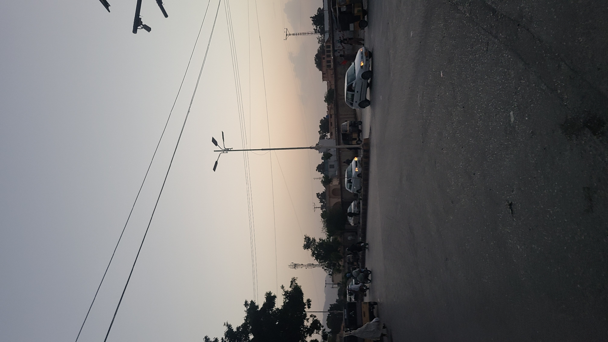 The image depicts a street scene in Quetta City Tehsil, Pakistan. The scene is taken at dusk, with a pale sky. The road occupies a significant portion of the lower right of the frame. Cars, scooters, and possibly other vehicles are on the road, some with headlights visible. Buildings and trees line the opposite side of the road. A utility pole and wires are on the left side of the frame.