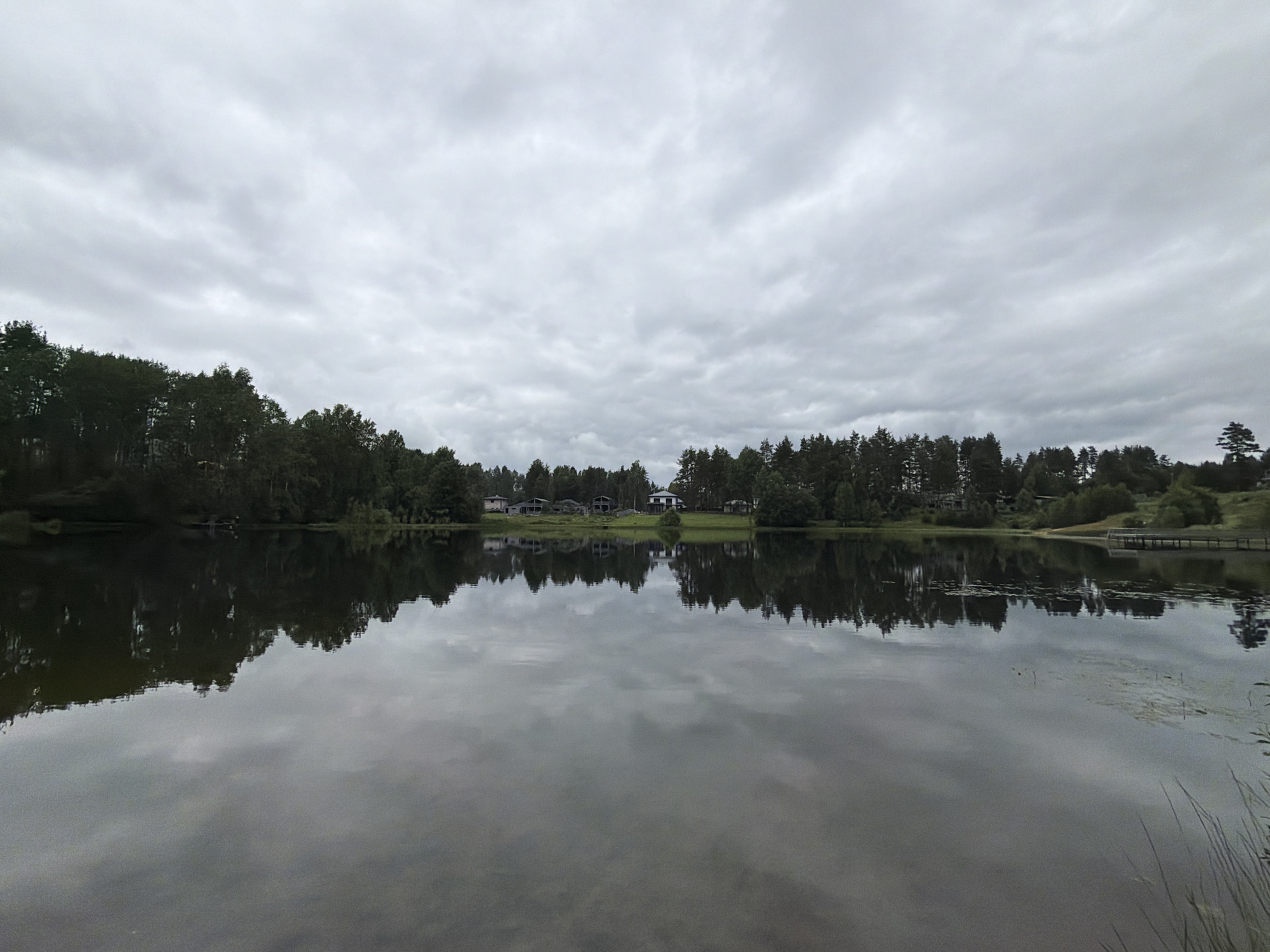 The image shows a body of water, likely a lake or large pond, reflecting a cloudy sky and a treeline along its edge. On the opposite shore, a few buildings, possibly houses, are visible amidst dense trees. The water's surface is calm, creating a clear reflection of the sky and the vegetation. To the right of the frame, a wooden pier extends into the water. The overall atmosphere is serene and overcast. The location is identified as Vaskelovo, Russia.