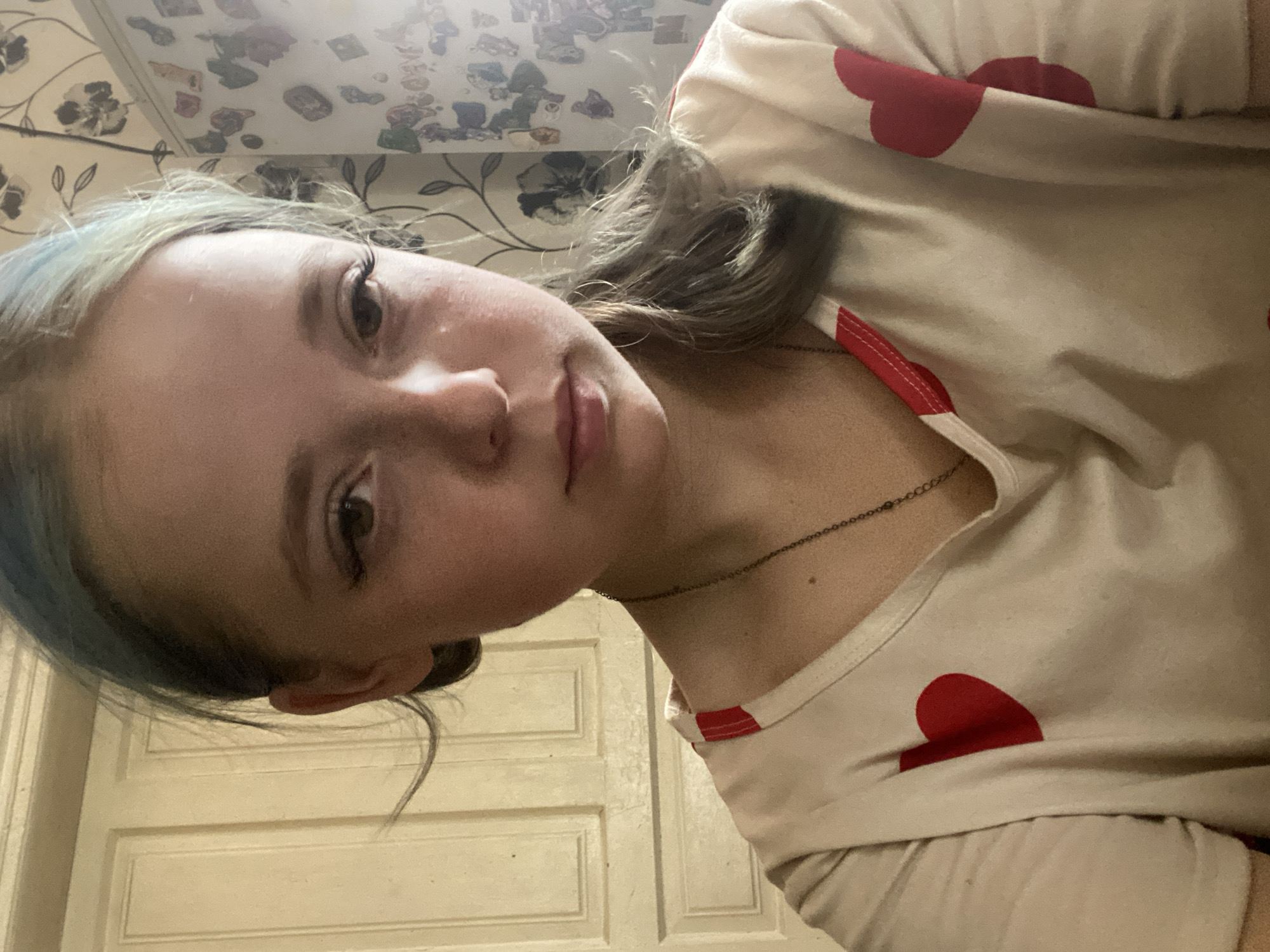 This media file features a close-up, low-angle shot of a young person, possibly a girl, with fair skin and light brown hair with hints of blue. She is looking towards the camera, her face slightly turned to the side. Her eyes are open, and she appears to have makeup on, including mascara. She is wearing a light-colored t-shirt with a repeating pattern of red hearts. A thin, dark necklace is visible around her neck.
The background consists of a textured wall with a floral pattern and what appears to be a door with paneling. The lighting suggests an indoor setting, possibly during the day given the brightness. There is no visible text in the image. The overall impression is a casual, personal snapshot. Given the provided location context of Nova Praha, Ukraine, the scene is likely within a residential interior in that area. No specific activity is clearly depicted; the subject is posing for the photo. The emotion conveyed is neutral to slightly inquisitive due to her gaze.