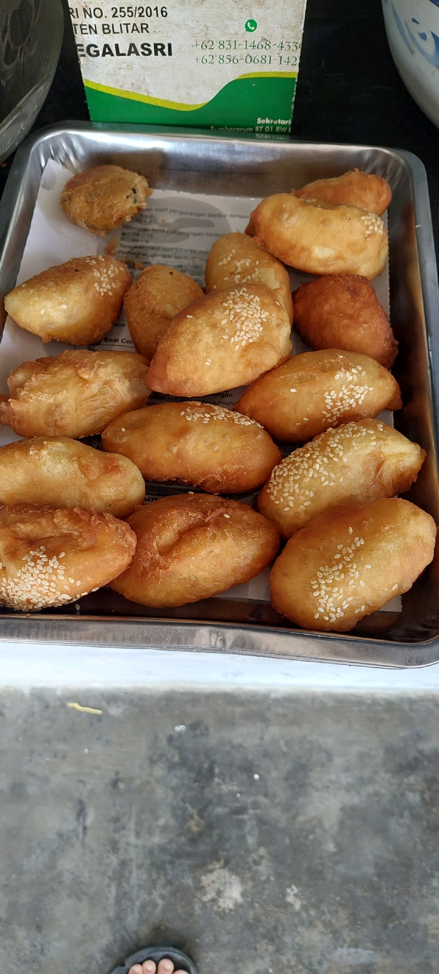 A tray of golden-brown fried pastries, some sprinkled with white sesame seeds, is displayed on a silver metal tray. The tray is placed on a surface that appears to be a concrete floor. In the background, a portion of a white and green box with text, including a phone number, is visible. The pastries vary slightly in shape and size, suggesting they are handmade. The overall impression is one of a food presentation, likely for sale or sampling.