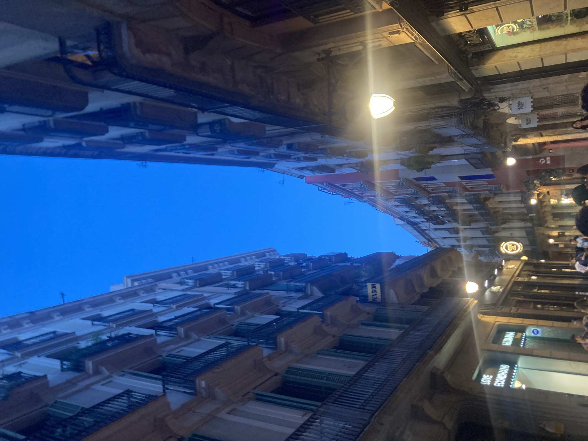 The image depicts a narrow street in Barcelona, Spain, at dusk. Tall, old buildings with balconies line both sides of the street, which is illuminated by streetlights and the light from shops. A blue twilight sky is visible above. On the left side of the street, a sign for "LOVE Econdhand" and "HUMANA" is visible on a shop. Further down on the left, a yellow sign reads "PENSION". On the right side of the street, shops also display illuminated signs, and a streetlamp hangs overhead. Several people are walking in the street, some appearing to be tourists. The architecture suggests an older district, possibly the Gothic Quarter or El Born.