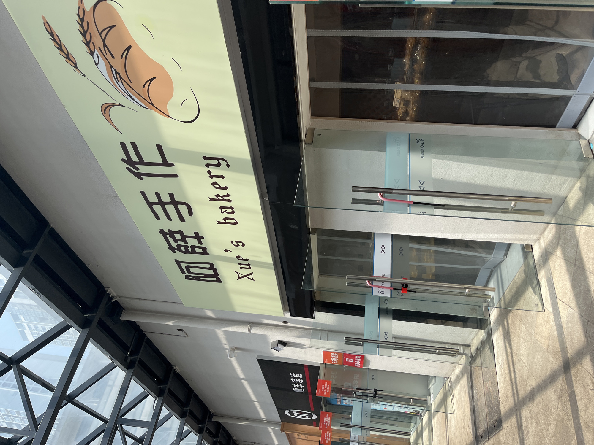 This image captures a bright, modern commercial interior, likely a shopping mall or a retail complex, located in Yuzhong District, China. The primary subject is the storefront of a bakery named "Xue's bakery."
The bakery's light green sign, oriented vertically due to the photo angle, features an illustration of a golden loaf of bread accompanied by wheat stalks. Below the illustration, the English text "Xue's bakery" is clearly visible, alongside Chinese characters "血旺手作?" which likely translates to "Xue's Handmade" or a similar phrase, despite the literal meaning of "血旺" (blood curd).
The storefront is composed of clear glass doors with sleek metal handles, reflecting the bright environment. The reflections show elements of the opposite side of the hallway, including other glass partitions and a glimpse of what appears to be another store or structure. Strong shadows are cast on the light-colored tiled floor, suggesting abundant natural light, possibly from skylights or large windows, indicating it is daytime.
Further down the hallway, other commercial spaces with glass fronts are visible. Text detected by OCR, such as "32" on a red sign and "夥多站" (meaning "many partners station" or "group station"), can be seen on these other signs or in reflections. The building's architecture is modern, characterized by dark, geometric metal beams forming part of the overhead structure or an adjacent wall. No people or specific activities are depicted, focusing instead on the architectural and commercial aspects of the location.
