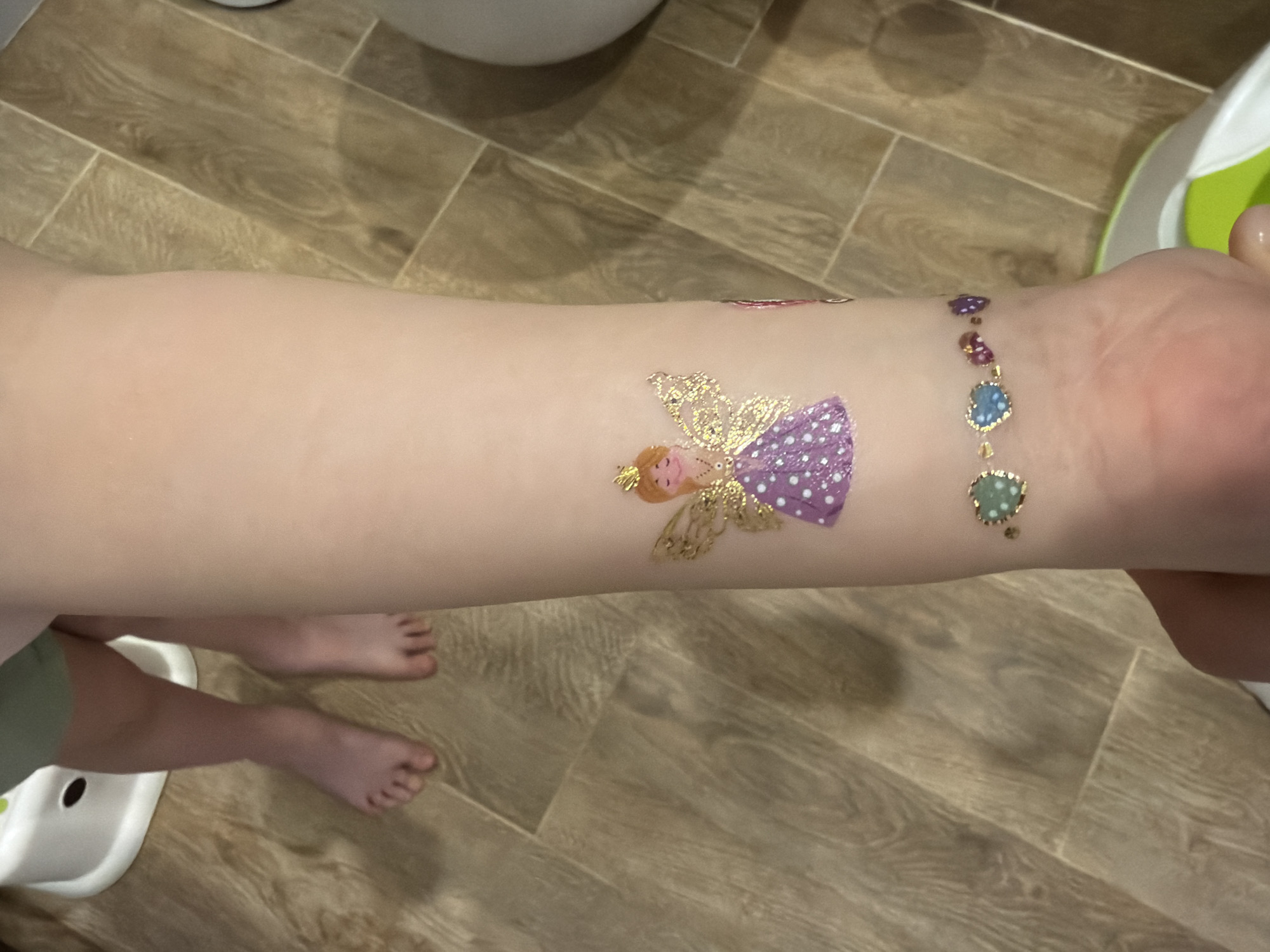 A close-up indoor photograph from Chaikovsky, Russia, captures a child's arm adorned with colorful temporary tattoos. The main subject is the child's left arm, held by an adult's hand on the right side of the frame. On the forearm, there is a glittery temporary tattoo depicting a fairy with golden wings and a purple dress with white polka dots. Further towards the wrist, another temporary tattoo creates the illusion of a bracelet, featuring a chain design with oval-shaped jewel-like elements in shades of purple, blue, and green, also with a glittery finish.
In the bottom left of the image, parts of a child's bare feet and lower legs are visible, resting on a light brown, wood-effect tiled or laminated floor. A white and green child's potty or toilet training seat is partially seen next to the child's feet. The background shows more of the wooden flooring, suggesting a domestic setting like a bathroom or child's play area. The lighting indicates an indoor daytime scene. No visible text is present in the image.
