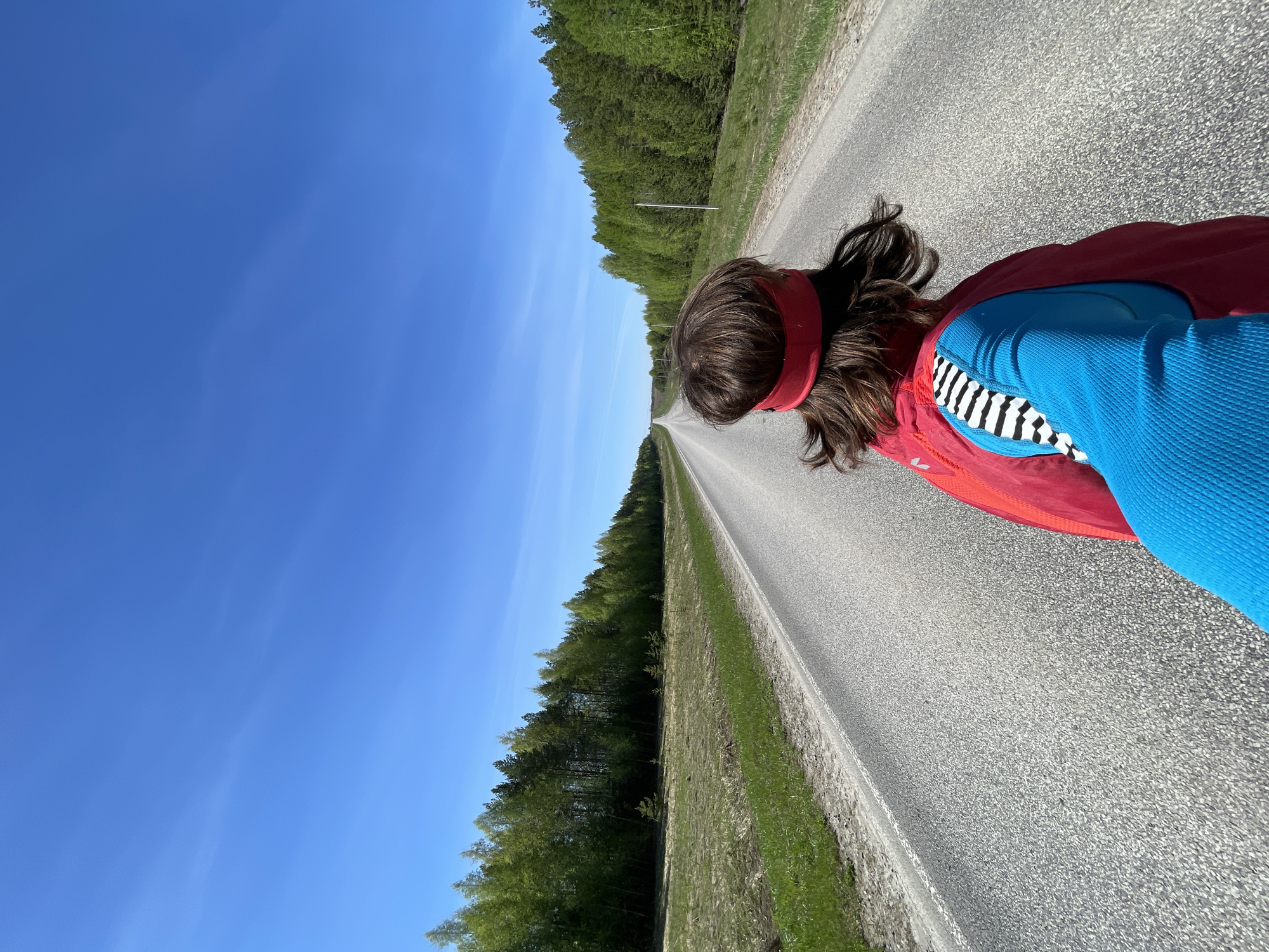 A person wearing a red headband, blue shirt, red jacket, and black and white striped undershirt is standing on an asphalt road. The road stretches into the distance, flanked by green trees on both sides. The sky above is clear and blue. The location is Перевозное, Russia.