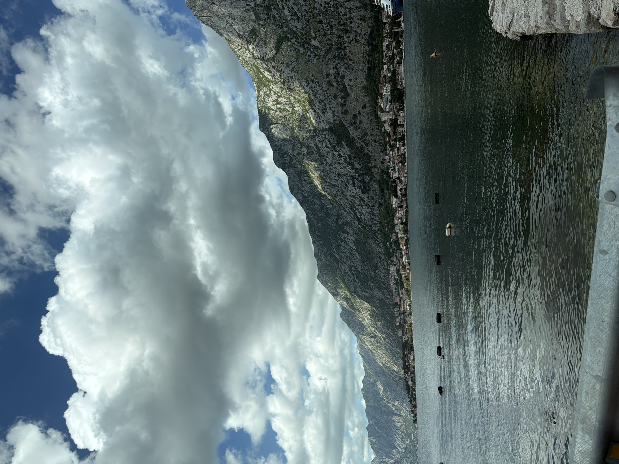 The image captures a view in Škaljari, Montenegro. A large, rocky mountain dominates the view, its slope dotted with sparse vegetation and urban development. The water, reflecting the sky's light, occupies a significant portion of the frame. A series of buoys are visible in the water. The sky is filled with fluffy white clouds against a backdrop of blue. Part of a barrier is present in the bottom right corner of the image.