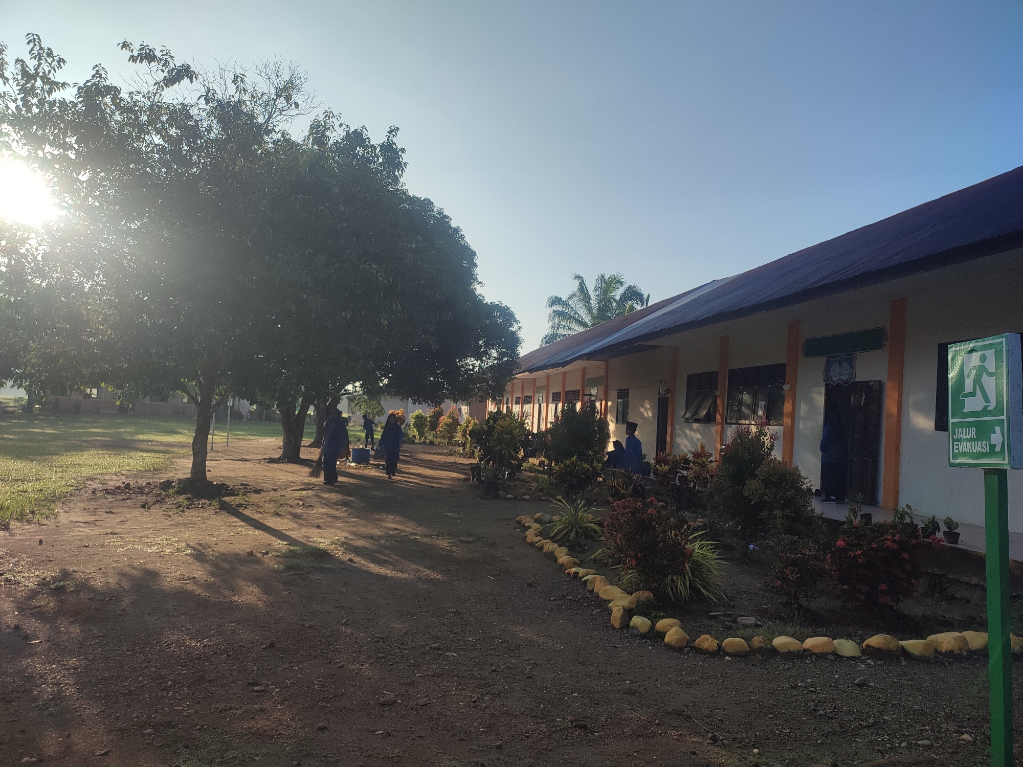 The image depicts an outdoor scene in Blangriek, Indonesia, with a low-slung building on the right side of the frame. The building appears to be a school or similar institution, with multiple windows and an overhanging roof. A green sign with a white figure running and the words "JALUR EVAKUASI" is positioned prominently near the building's entrance. In front of the building, there is a small garden area delineated by a border of rocks. Several people wearing blue uniforms are visible along a dirt path leading away from the building. To the left, a large tree obscures the sun, creating dappled shadows on the ground. The ground is primarily dirt, with patches of grass in some areas. The sky is clear and light blue.