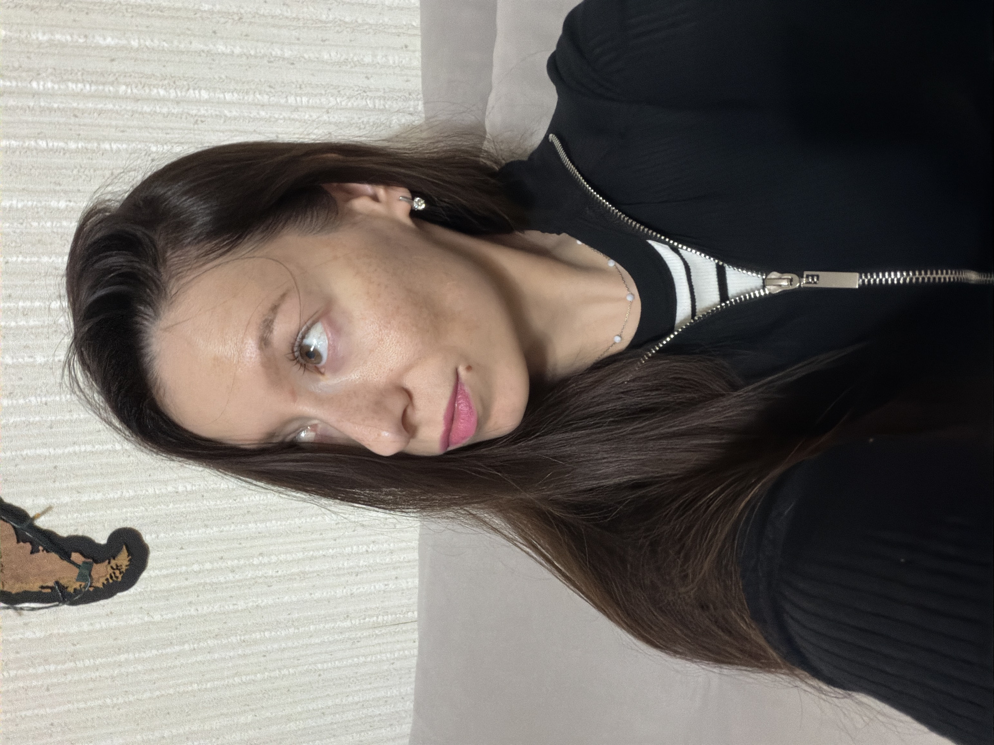 A woman with long, dark brown hair, fair skin with freckles, and pink lipstick is shown from the chest up, looking to her left. She is wearing a black zippered jacket over a black and white striped shirt. A delicate necklace with light-colored beads is visible around her neck, and she has a small diamond earring in her left ear. The background consists of a textured, light-colored wall with a decorative item resembling a butterfly on the upper left. The scene appears to be indoors, possibly in a living room, as a light-colored couch is partially visible behind her. There is no indication of the city of Raevka, Russia.