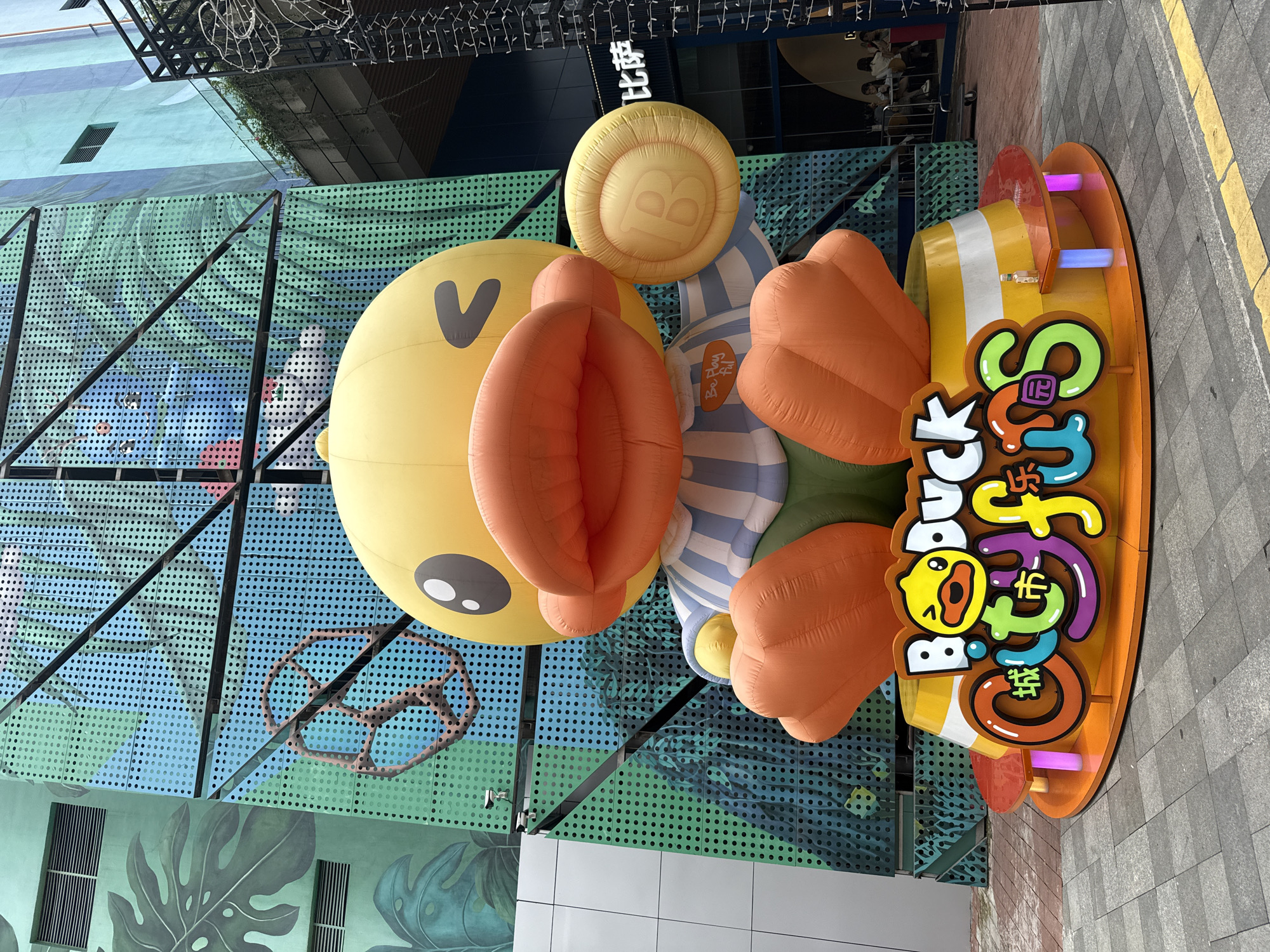 In Huizhou, China, an enormous inflatable yellow duck character is prominently displayed as an outdoor art installation. The duck, adorned with a blue and white striped shirt and green shorts, sits on a circular, orange and yellow base featuring the words "Bo-duck City Fun" in colorful, stylized English and Chinese characters. The duck is winking and holding a round, yellow object with the letter "B" embossed on it in its right hand. Behind the duck, a building facade is decorated with a large-scale mural featuring tropical foliage in shades of green and teal, interspersed with a grid pattern of small, circular openings. The overall scene appears to be in an urban or commercial area, with paved ground in the foreground and hints of other structures and lighting in the background. The lighting suggests it is daytime.