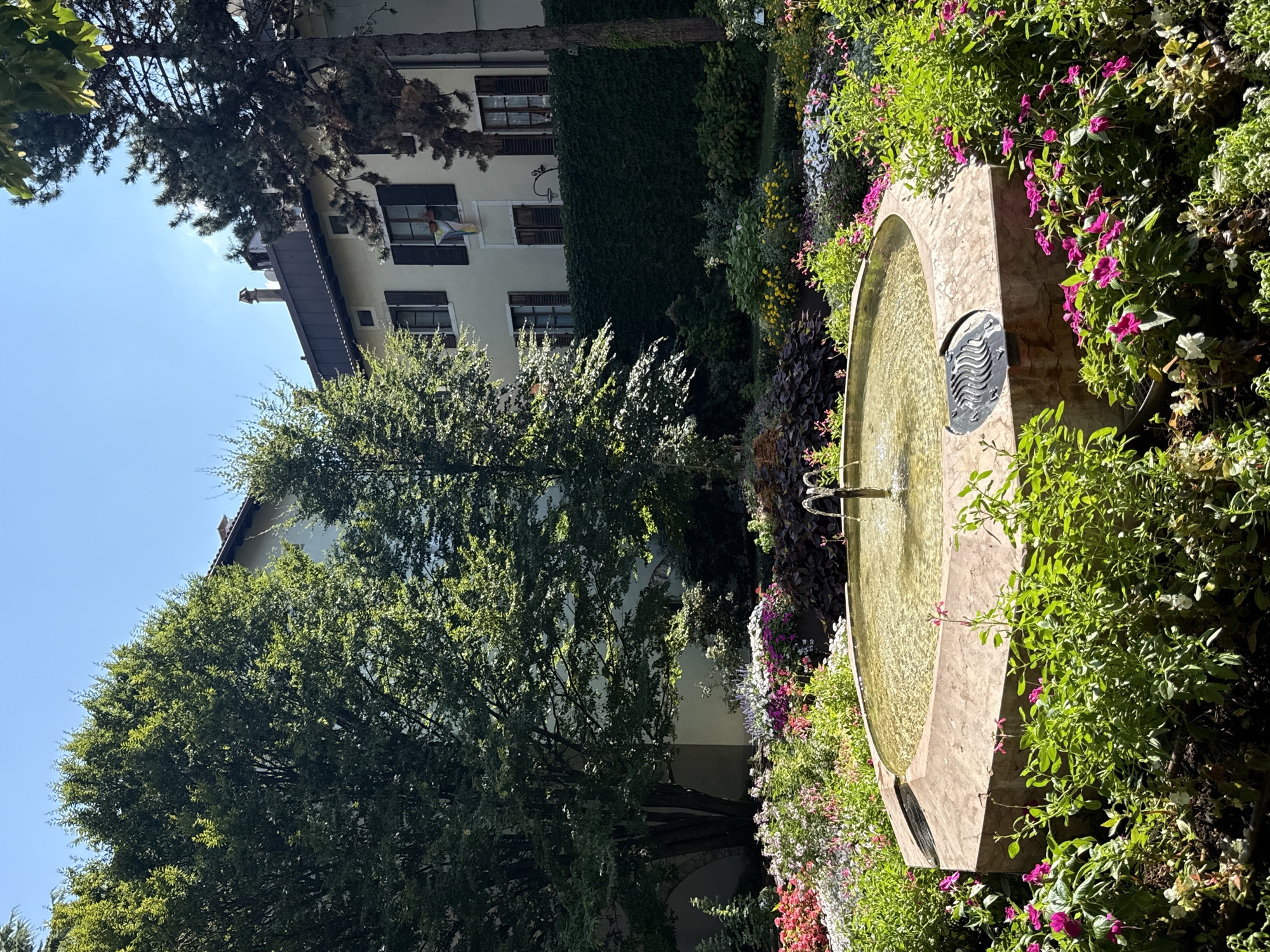 A circular, light-brown fountain with a marbled texture is centrally located in a lush garden in Trento, Italy. The fountain is filled with clear water, and a thin stream of water shoots up from its center. The fountain is surrounded by a vibrant array of flowers in various colors, including pink, white, and purple, with abundant green foliage. In the background, a two-story building with a light-colored facade and dark shutters is visible. The building has a dark-tiled roof and a chimney. Several mature trees with dense green foliage are scattered throughout the scene, casting shadows on the ground. The sky above is clear and blue. The overall impression is a tranquil and beautiful garden setting on a sunny day.