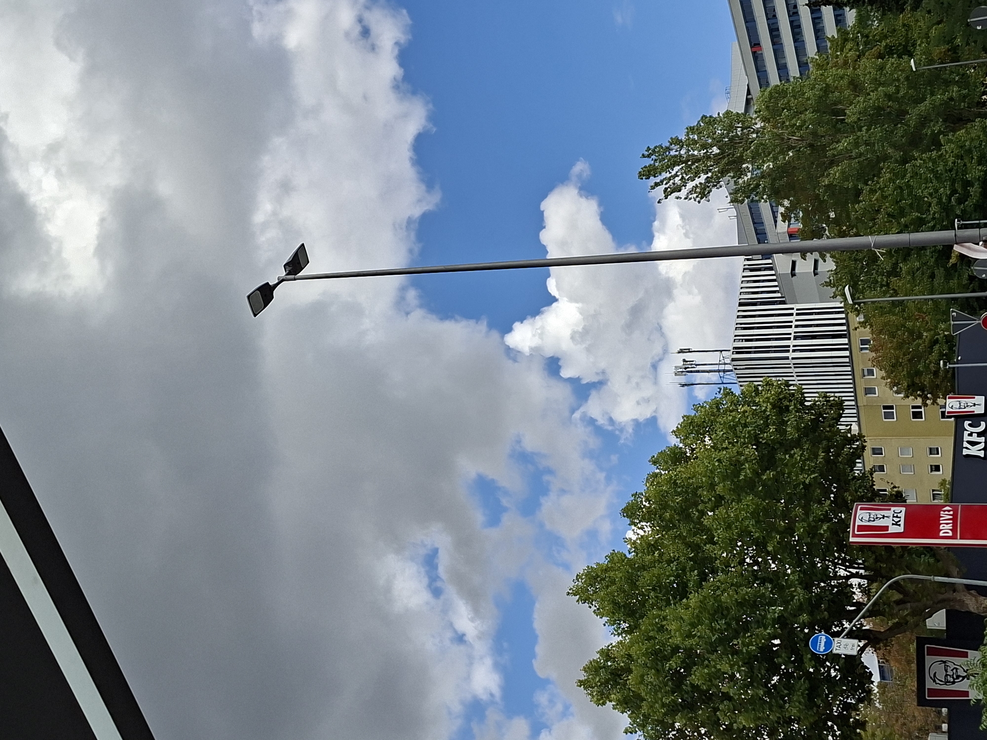 The image shows a street scene in Wiesbaden, Germany. A tall, grey lamppost with two lights stands prominently in the foreground, reaching towards a cloudy sky. To the left, a large, leafy green tree dominates the frame. Behind the tree, there is a KFC restaurant with its characteristic red and white signage, including a "DRIVE-THRU" sign. Further back, a modern building with a black and white striped facade is visible, along with other apartment or office buildings in the background. The sky is a mix of blue and grey clouds, suggesting a partly cloudy day.