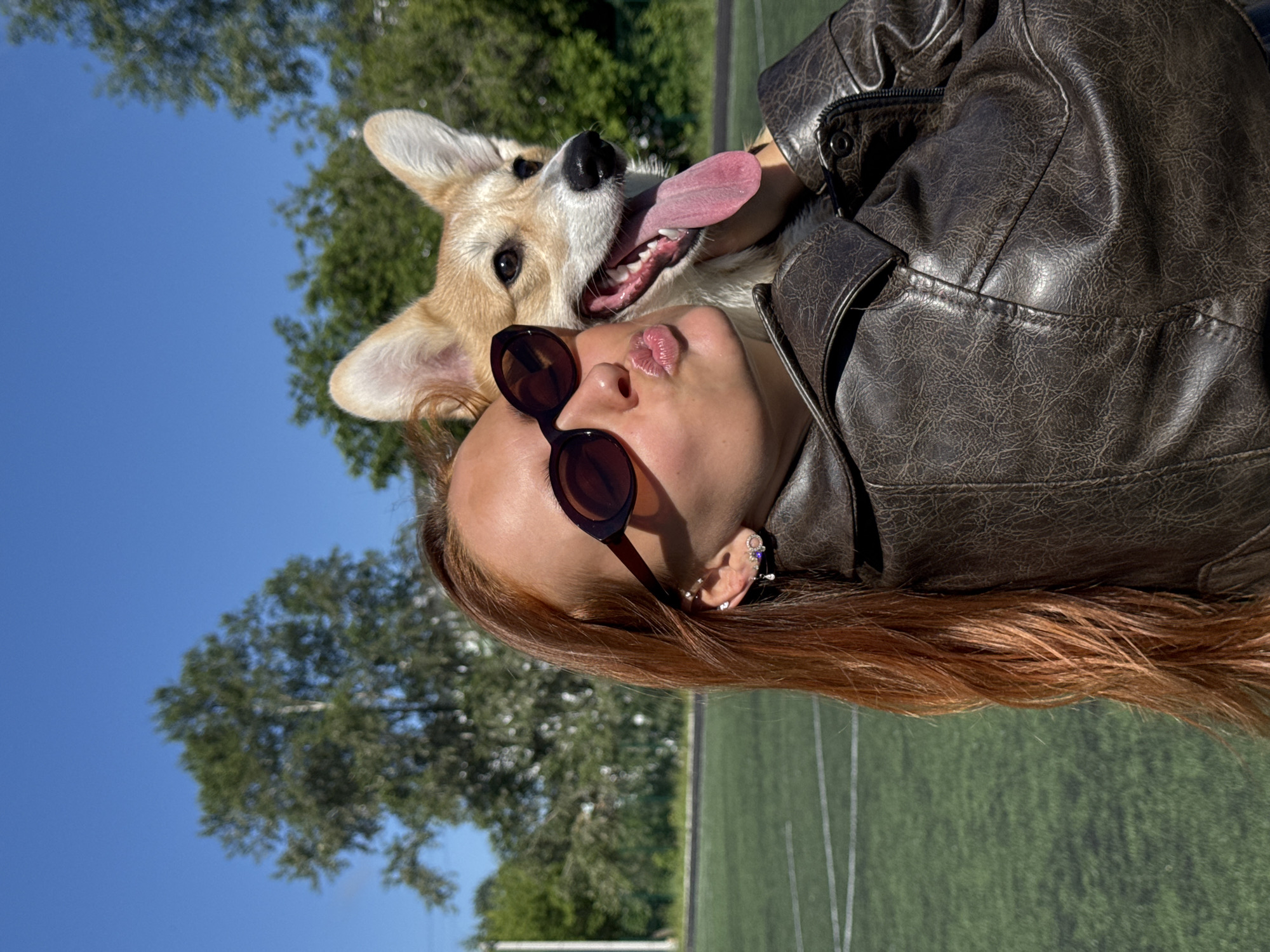 A young woman with long, auburn hair and sunglasses is taking a selfie with a corgi dog outdoors on a sunny day. She is wearing a dark brown leather jacket and making a kissing face. The corgi has its tongue out, appearing happy. They are standing on a green sports field with white markings, possibly a soccer field, with trees and a blue sky in the background. The location is a park or sports facility in the city of Chaikovsky, Russia.