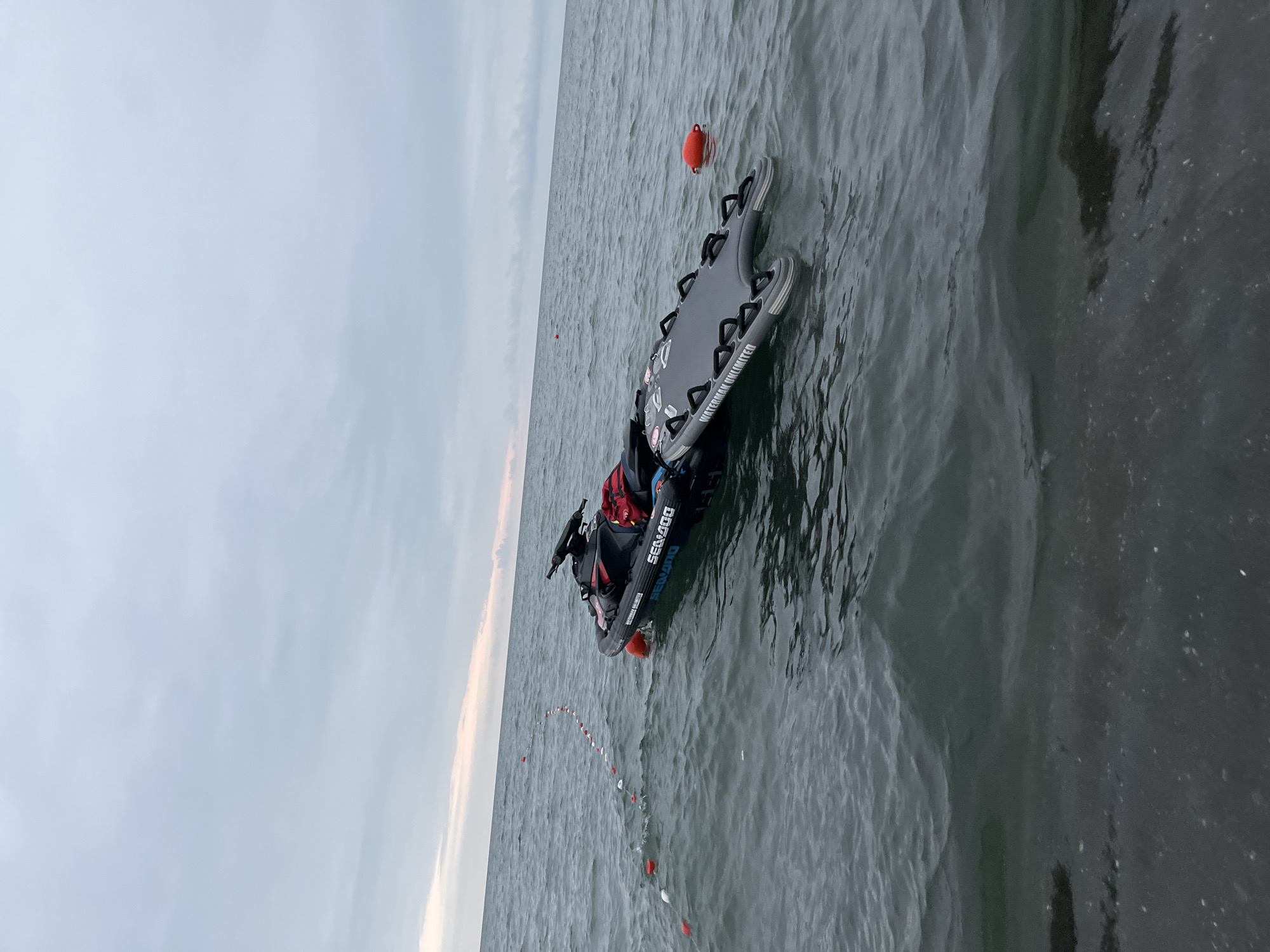 The image depicts a jet ski with a rescue board attached, floating on the water near the shore in Ureki, Georgia. The sky is overcast with muted, soft colors, suggesting either early morning or late evening. The water is calm with gentle ripples. A rope with buoys stretches across the water to the left of the jet ski, marking a boundary. The jet ski itself is black with "SEADOO" branding and appears to be a model designed for rescue operations. A red bag is secured to the back of the jet ski. The rescue board, labeled "WATERMAN UNLIMITED," is grey and strapped to the jet ski. A single orange buoy is visible in the water to the right of the jet ski. The shoreline is visible in the foreground, appearing dark and wet, with some small debris or foam on the water's surface near the edge. The scene suggests a prepared state for water rescue or training exercises.