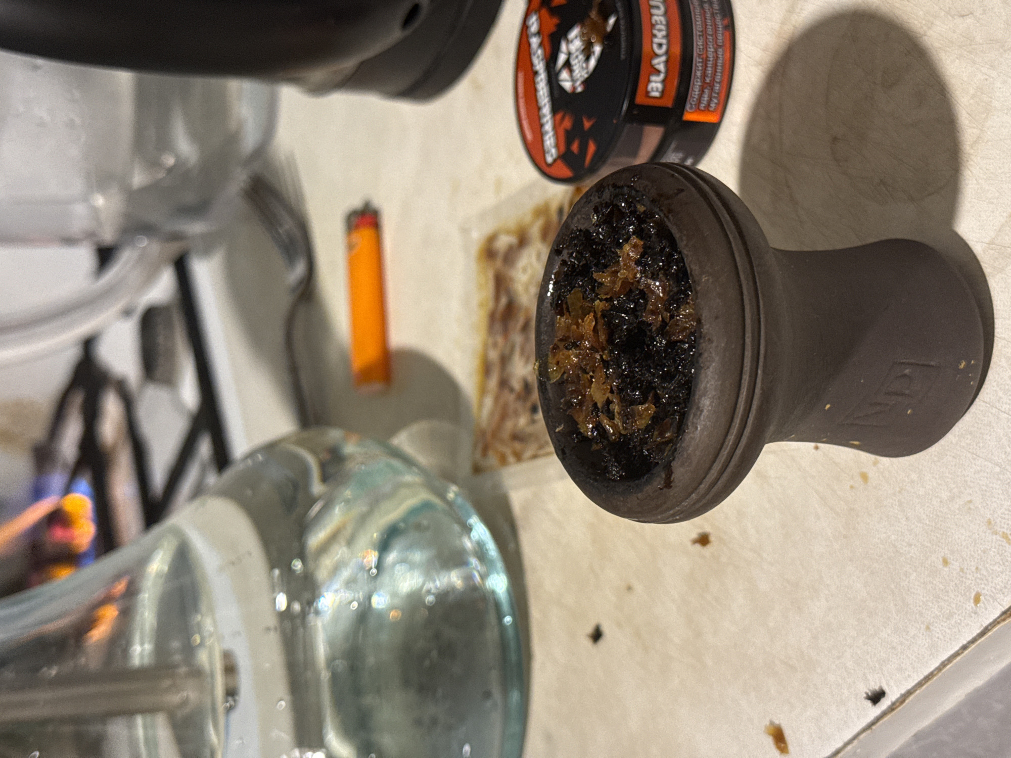 The image shows a hookah setup on a light-colored surface, possibly a table or countertop. The hookah includes a glass base, a metal stem, and a ceramic bowl filled with dark, possibly burnt, hookah tobacco. Next to the bowl is an open package of shisha tobacco and a lighter. A can of "BLACK BURN" tobacco is also visible. Given the brand of tobacco, it is likely that the location is Moscow, Russia, as this is a Russian brand.