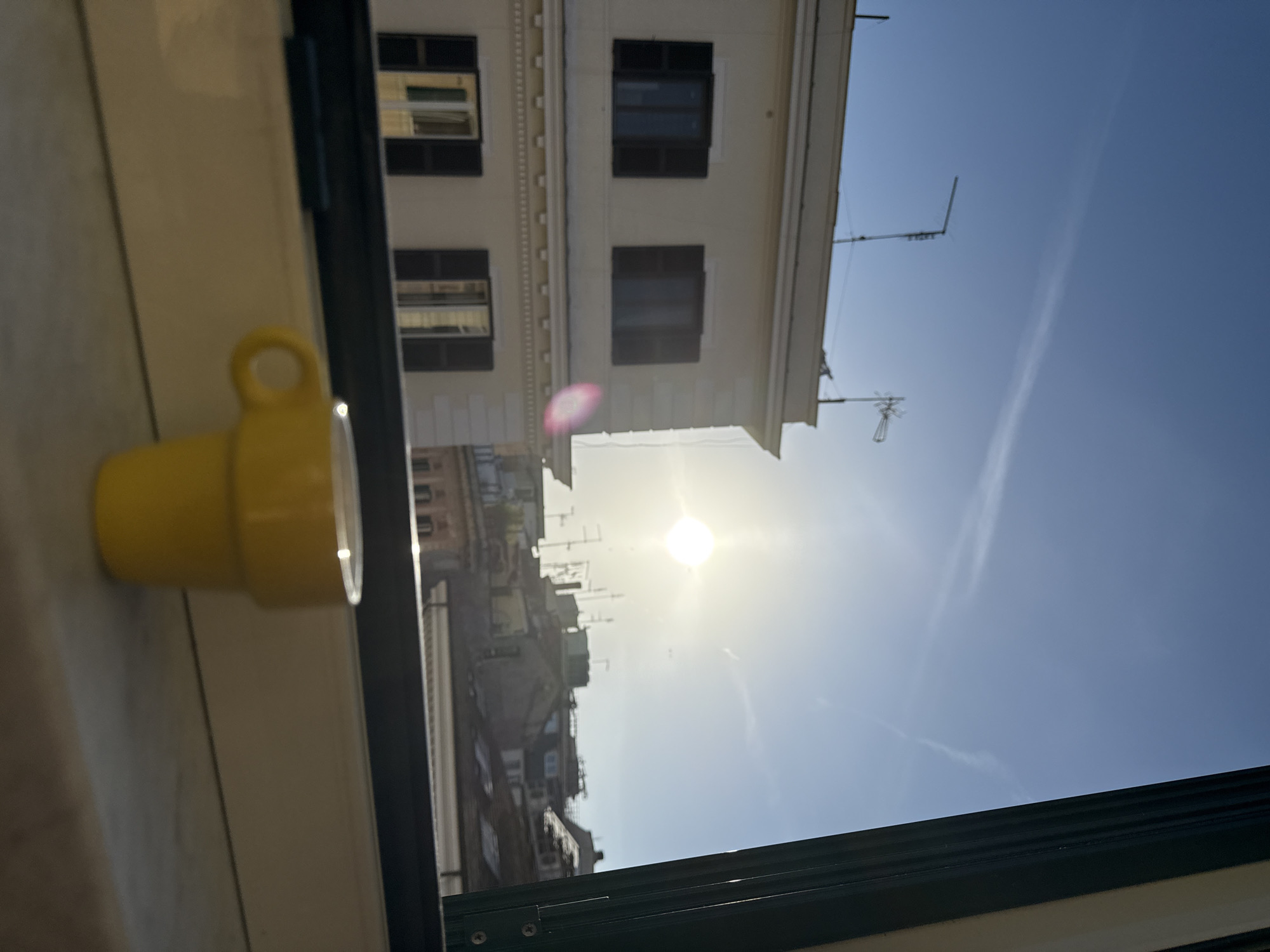 The image, captured in portrait orientation (originally rotated 90 degrees clockwise), presents a bright, sun-drenched urban view from inside a building. In the immediate foreground, a vibrant yellow ceramic mug with a handle rests on a light-colored window sill on the left side of the frame. The dark frame of the window borders the view on the right and bottom.
Beyond the window, a classic European cityscape unfolds under a clear blue sky. Directly across, a multi-story building with a light-colored, stucco-like facade features several rectangular windows, some fitted with closed dark shutters. Architectural details like cornices are visible. Further into the distance, a dense array of rooftops, additional buildings, and numerous television antennas or aerials create a textured urban fabric.
The sun is a brilliant, almost blinding orb in the sky, positioned towards the center-right of the outdoor scene, indicating a clear, sunny day, likely late morning or early afternoon. A faint white contrail stretches diagonally across the blue expanse of the sky. The architecture, particularly the building styles and roofline details, strongly suggests a Mediterranean or Southern European city, consistent with the given location context of Rome, Italy. The scene conveys a tranquil, observational mood from indoors. No specific human activity or visible text is present.