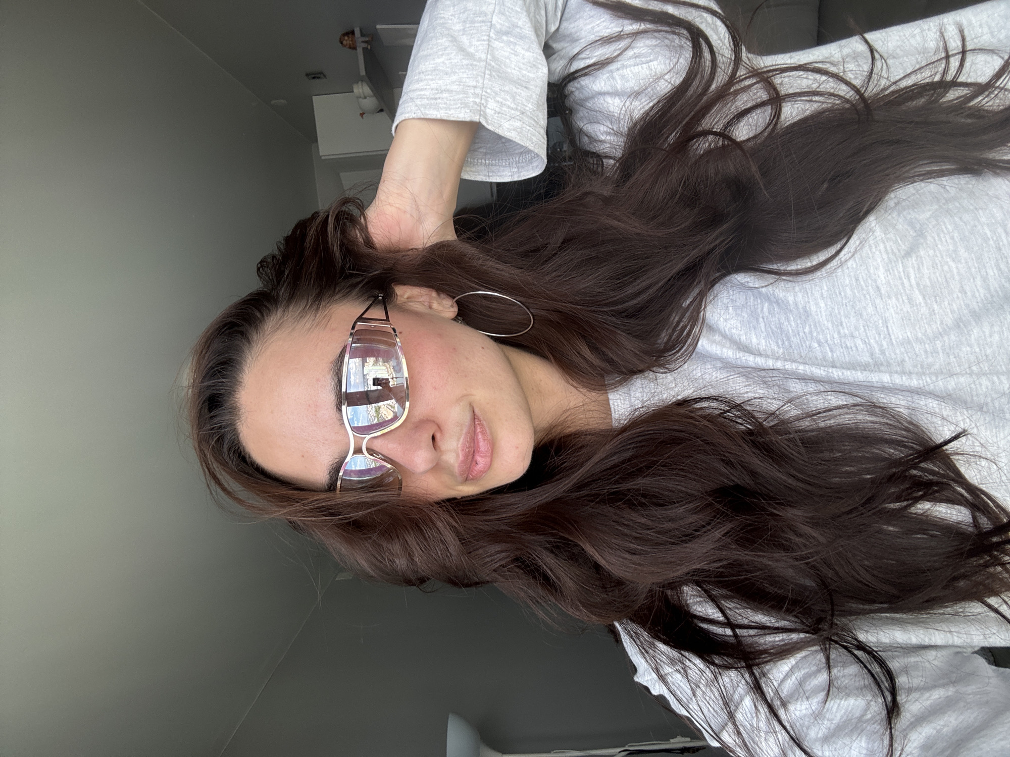 A woman with long, dark brown hair is seen from a close, slightly angled perspective. She's wearing wire-rimmed glasses reflecting a bright, possibly outdoor, scene and a light-colored, long-sleeved shirt. She is also wearing hoop earrings. Her hand is near her head, possibly holding her hair. The background consists of a gray wall and glimpses of a room interior, including what appears to be a white cabinet or furniture piece. In the reflection of the glasses, the cityscape visible appears to be Krasnoyarsk, Russia, indicated by the presence of structures resembling the Communal bridge, typical Siberian architecture, and the Yenisey River.