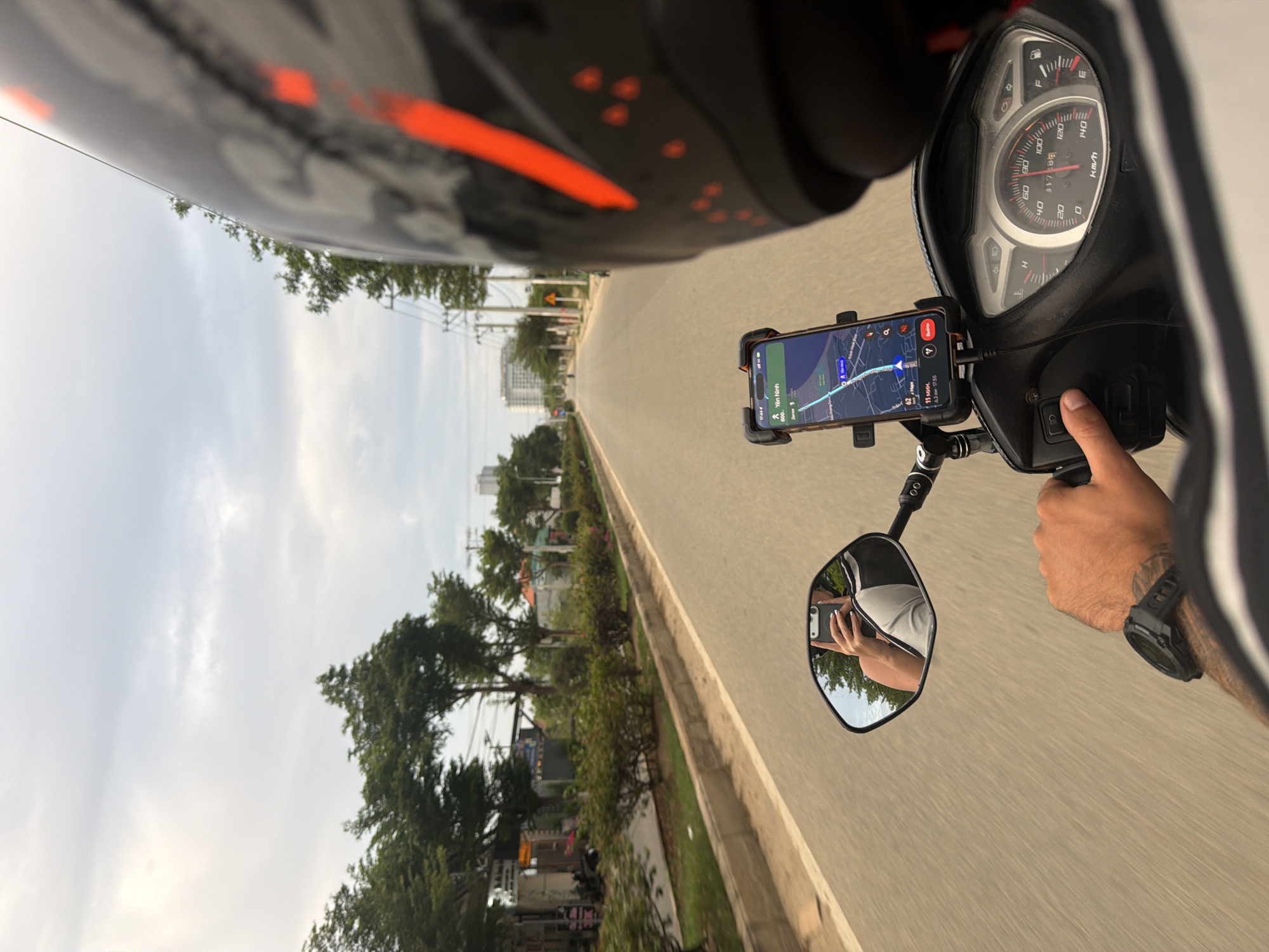 The image presents a first-person perspective from a scooter rider's point of view, looking forward down a road under an overcast sky. The rider's hand is visible on the right handlebar, clutching the grip, with a black digital watch on the wrist. A portion of the rider's helmet, black with orange accents, is visible at the top left.
Mounted centrally on the handlebars is a smartphone held in a cradle, displaying a navigation application. The app shows a map with a blue route line, indicating a destination labeled "Yên Ninh" and showing "11 min" travel time for "6.3 km," with an estimated arrival time of "17:55." The phone is connected via a charging cable to the scooter.
To the right of the phone, the scooter's instrument cluster is visible. It features a speedometer calibrated in "km/h," with markings up to 140. The needle rests at approximately 60 km/h, suggesting the scooter is in motion. A fuel gauge with "F" (full) and "E" (empty) indicators is also present, showing the tank is nearly full. An odometer reading of "31728" is discernible.
On the far right, a rearview mirror reflects the rider holding a smartphone, capturing the image. This indicates the photo is a selfie taken while riding. The road ahead is asphalt, with a clear median strip lined with greenery and small trees. Further in the distance, more trees and some taller buildings are visible, suggesting an urban or semi-urban setting. The sky is largely covered with light clouds, indicating an overcast day or late afternoon. Based on the navigation app's time, it is late afternoon in Dong Hai Ward, Vietnam.