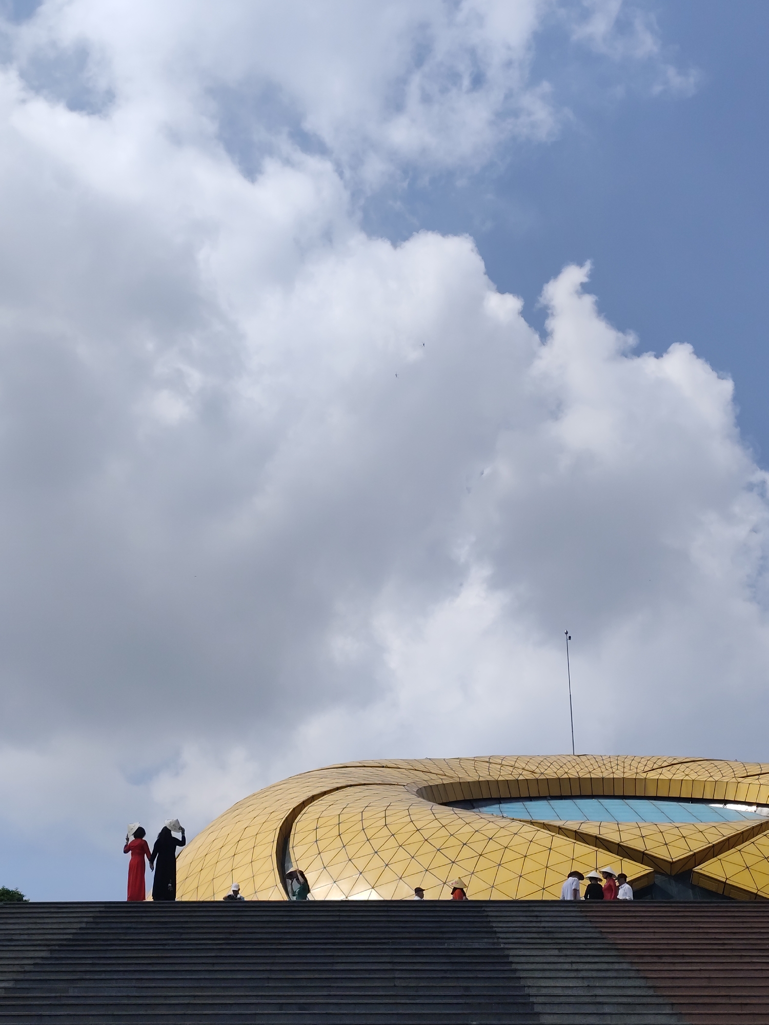 The photo depicts a partial exterior view of the Golden Dome in Da Lat, Vietnam, including a staircase leading to the dome. Two people in traditional Vietnamese clothing, one in red and one in black, stand on the stairs, each holding a white conical hat. Several other people stand on the stairs and near the base of the dome. The dome is gold-colored with geometric patterns and a large circular opening showing a blue interior. A tall antenna rises from the top of the dome. The sky is partially cloudy with patches of blue sky visible. The lower portion of the image is dominated by the steps leading up to the dome.