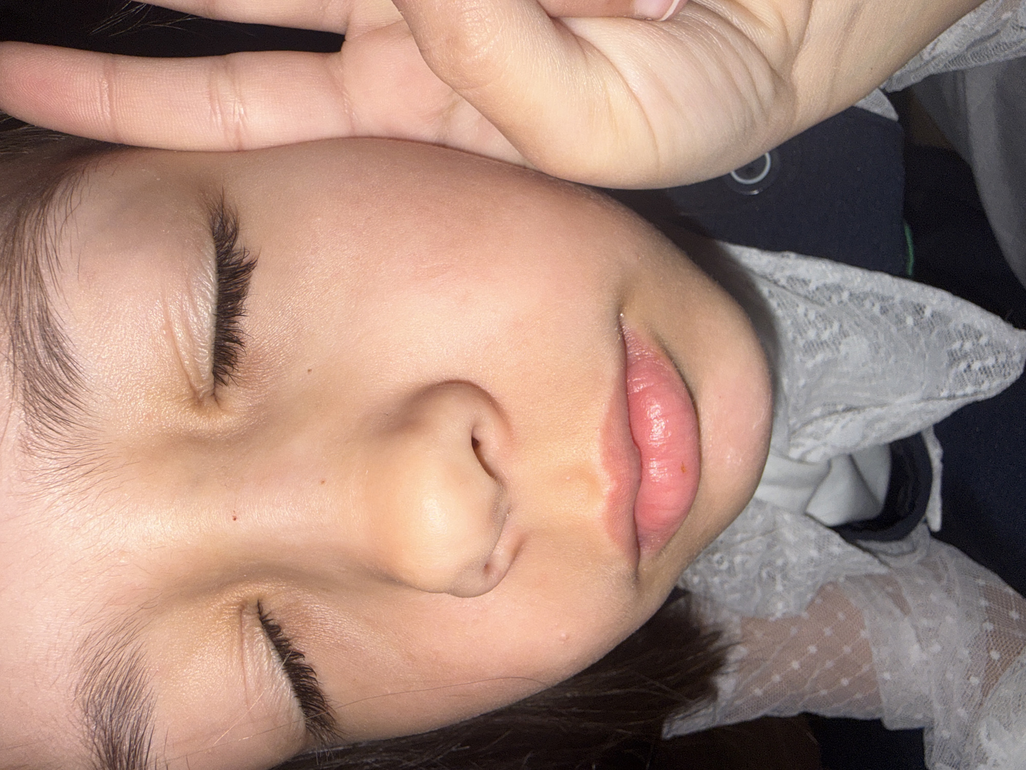 The image shows a close-up of a child's face in repose. The child's eyes are closed, and a hand is positioned above their forehead. The skin appears smooth and fair, with visible eyelashes and eyebrows. The child's lips are slightly parted. A white, lacy collar is visible on the right side of the frame. The background is dark and out of focus. There is no evidence in the image to suggest a specific location, including Votkinsk, Russia.