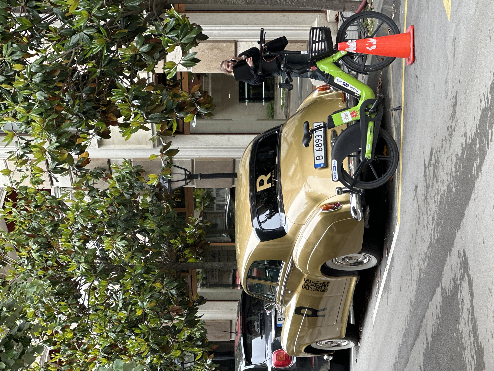 This daytime image, set in Varna, Bulgaria, depicts a street scene with a vintage car, an electric bicycle, and a person. A gold-colored vintage car, possibly a classic model, is prominently parked on the right. It features white-wall tires, chrome hubcaps, and is adorned with black text including "R" on the rear window, and "R.FITNESS" and a QR code on its side.
Adjacent to the car is a lime green shared electric bicycle (e-bike) with a front basket. A red traffic cone leans against the e-bike's front wheel. Standing beside the e-bike is a person, dressed in dark attire, holding what appears to be a black laptop or folder, and seemingly engaged in a phone call.
The ground surface is wet asphalt, suggesting recent rain. Yellow and white lines are painted on the street. On the left, a large tree with dense, dark green foliage, possibly a Magnolia, frames the scene and partially obscures the light-colored building facades in the background. A street light pole is also visible near the tree.
Visible text includes "R.FITNESS" on the vintage car. On the e-bike, "5 min. free daily" and a tag displaying "B 6893 TA" are present.