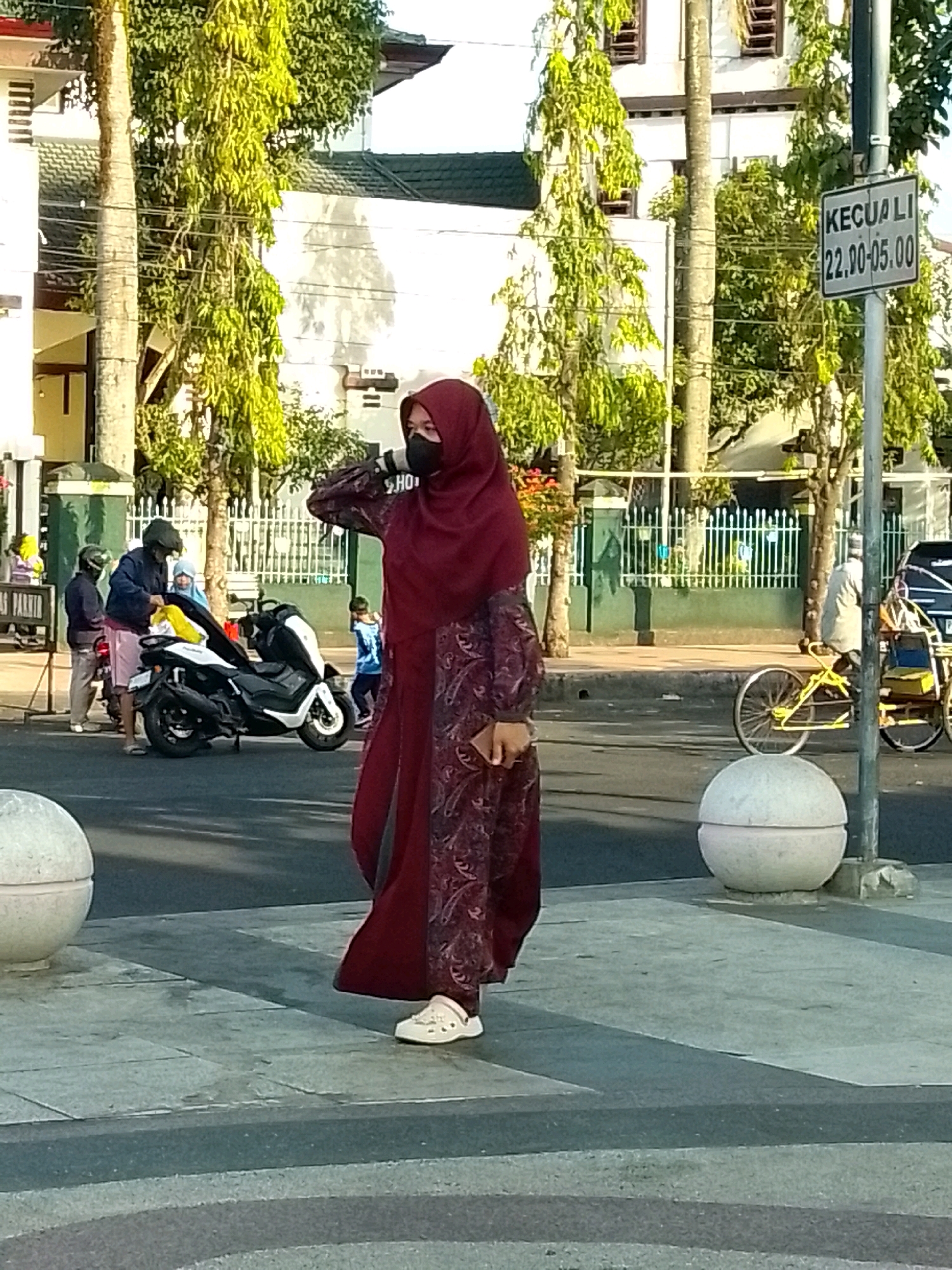 A full-body shot captures a woman wearing a maroon hijab and a matching patterned dress, standing on a sidewalk in Jember, Indonesia. Her right hand is raised to her head, touching the hijab, and she appears to be looking to her left. She is wearing white clogs. In the background, a motorcycle is parked on the street, with several people standing nearby. Further back, a rickshaw is visible, and a traffic sign with Indonesian text and numbers "22.00-05.00" is mounted on a pole. The scene is set against a backdrop of trees and buildings, suggesting an urban environment in the afternoon.