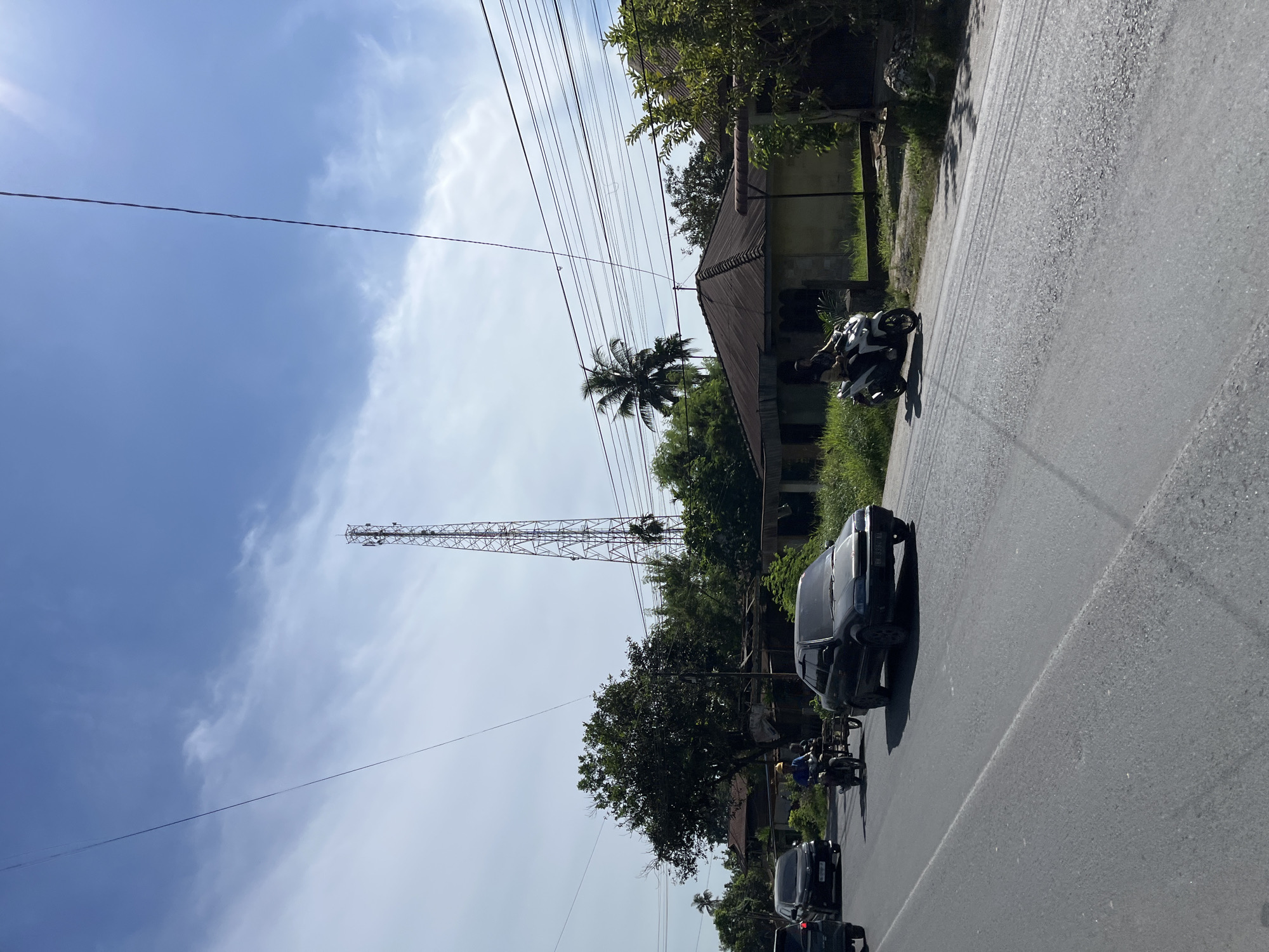 The image depicts a street scene in Bukitrata, Indonesia, under a bright, partly cloudy sky, suggesting it is daytime. The perspective is from a moving vehicle on the right side of the road.
The main subjects are vehicles on the road and a tall telecommunications tower in the background. On the road, a dark-colored van is prominent in the foreground, followed by a motorcycle. Further down the road, another black car and a few more motorcycles are visible. A person is riding the motorcycle in the foreground.
The setting includes a paved road with a white line marking its center. To the left of the road, there's a grassy area with trees and vegetation. A building with a corrugated metal roof and faded yellow walls is situated behind the grassy area. In the distance, a very tall, lattice-like telecommunications tower rises against the sky, with numerous power lines running horizontally across the scene. A single palm tree stands near the building.
The activity appears to be normal daily traffic on a road in a tropical or subtropical area. There are no strong emotional cues visible; the scene is one of ordinary daily life. No visible text is present in the image.