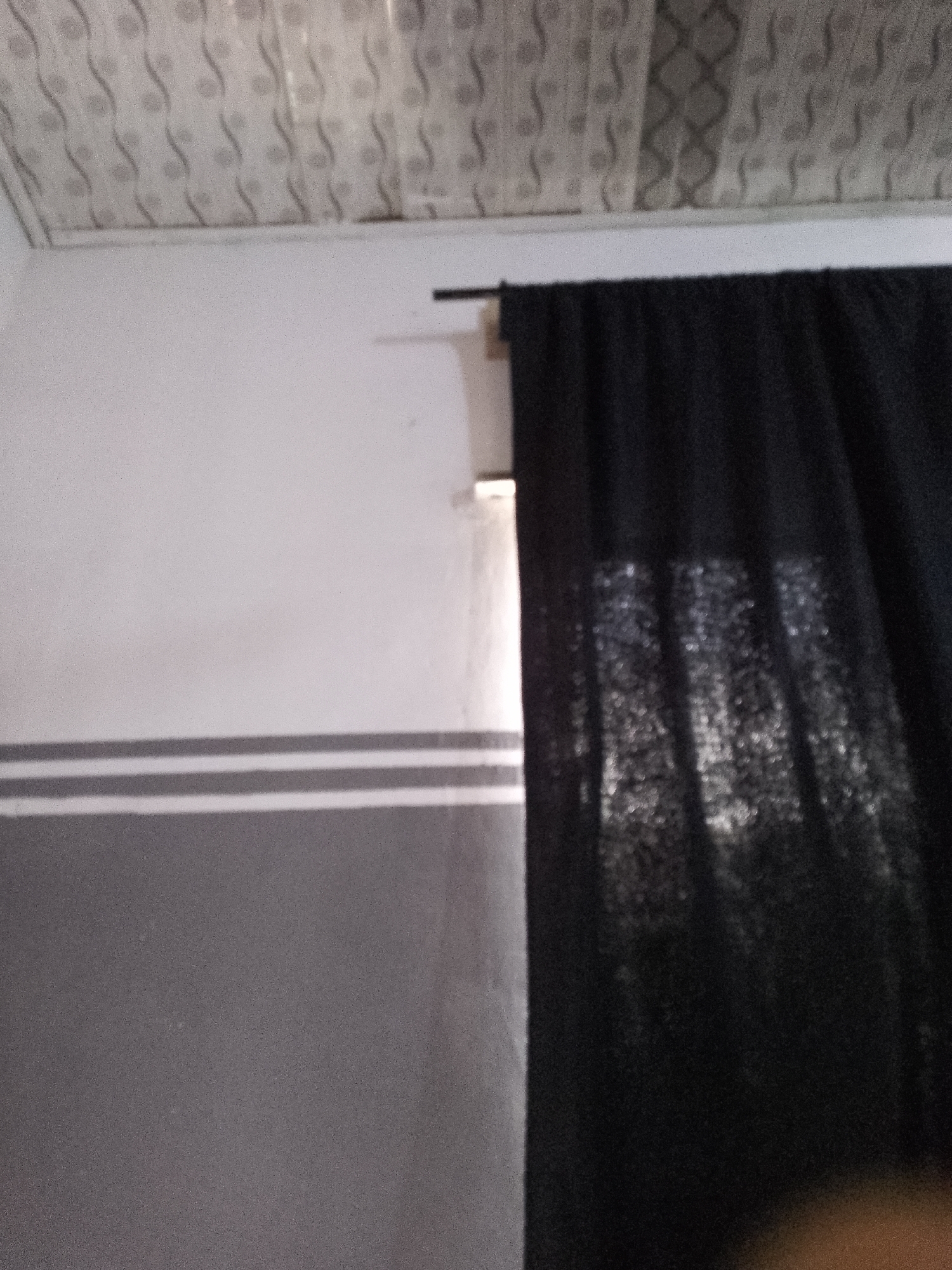 The image shows a room interior with a white ceiling, a grey and white striped wall, and a black curtain partially covering a window. The ceiling has a textured pattern. The wall is divided horizontally into two sections, with the upper section being white and the lower section grey. Two thin white stripes run across the grey section of the wall. The black curtain is drawn, allowing some light to filter through, creating a pattern of light and shadow on the fabric. The scene appears to be in Jalingo, Nigeria, based on the provided context. No people or specific actions are visible in the image.