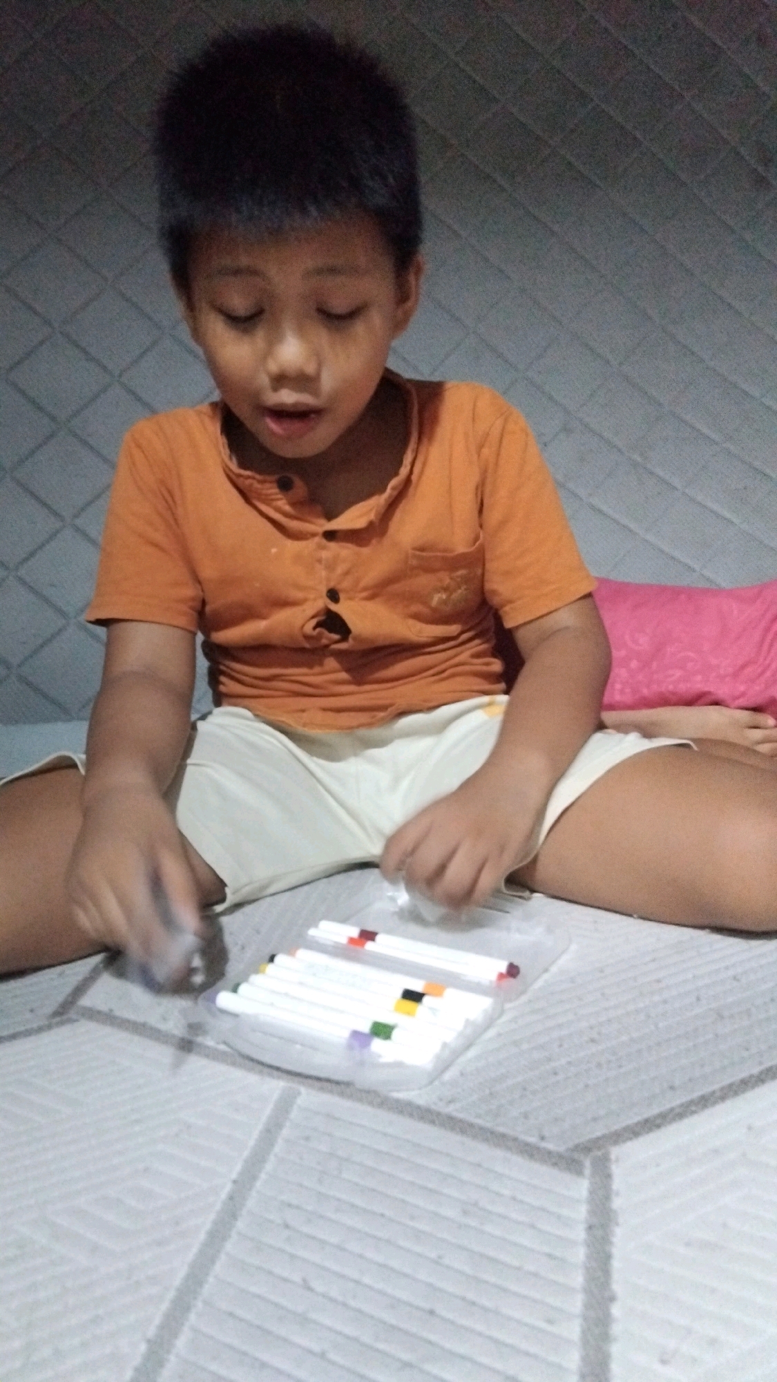 A young boy is sitting on a bed, looking down at a set of markers. He is wearing an orange t-shirt and light-colored shorts. The bed has a textured gray and white patterned mattress, and a pink pillow is visible on the right side of the image. The boy's eyes are closed, and his mouth is slightly open, suggesting concentration or being in the process of speaking. His hands are positioned in front of him, with one hand near the markers and the other blurred due to motion. The markers are neatly arranged in a clear plastic case, and various colors are visible on their caps. The scene appears to be indoors, with the background showing a patterned wall. The lighting suggests it could be daytime, but there are no clear indicators of time of day or weather. The overall mood seems to be one of focused activity and possibly creativity. There is no visible text in the image. The location context provided is Bangun Rejo, Indonesia.