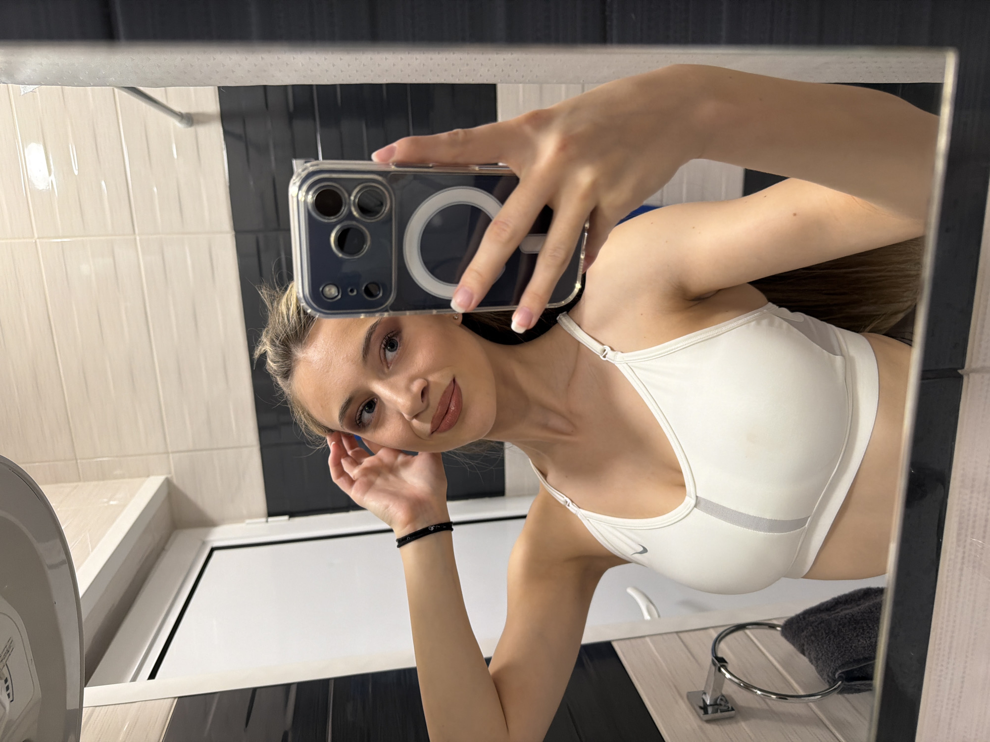 A woman takes a selfie in a bathroom. The bathroom has black and white striped tiles. There is a sink and a towel rack visible. The location is not discernible as Plovdiv, Bulgaria.