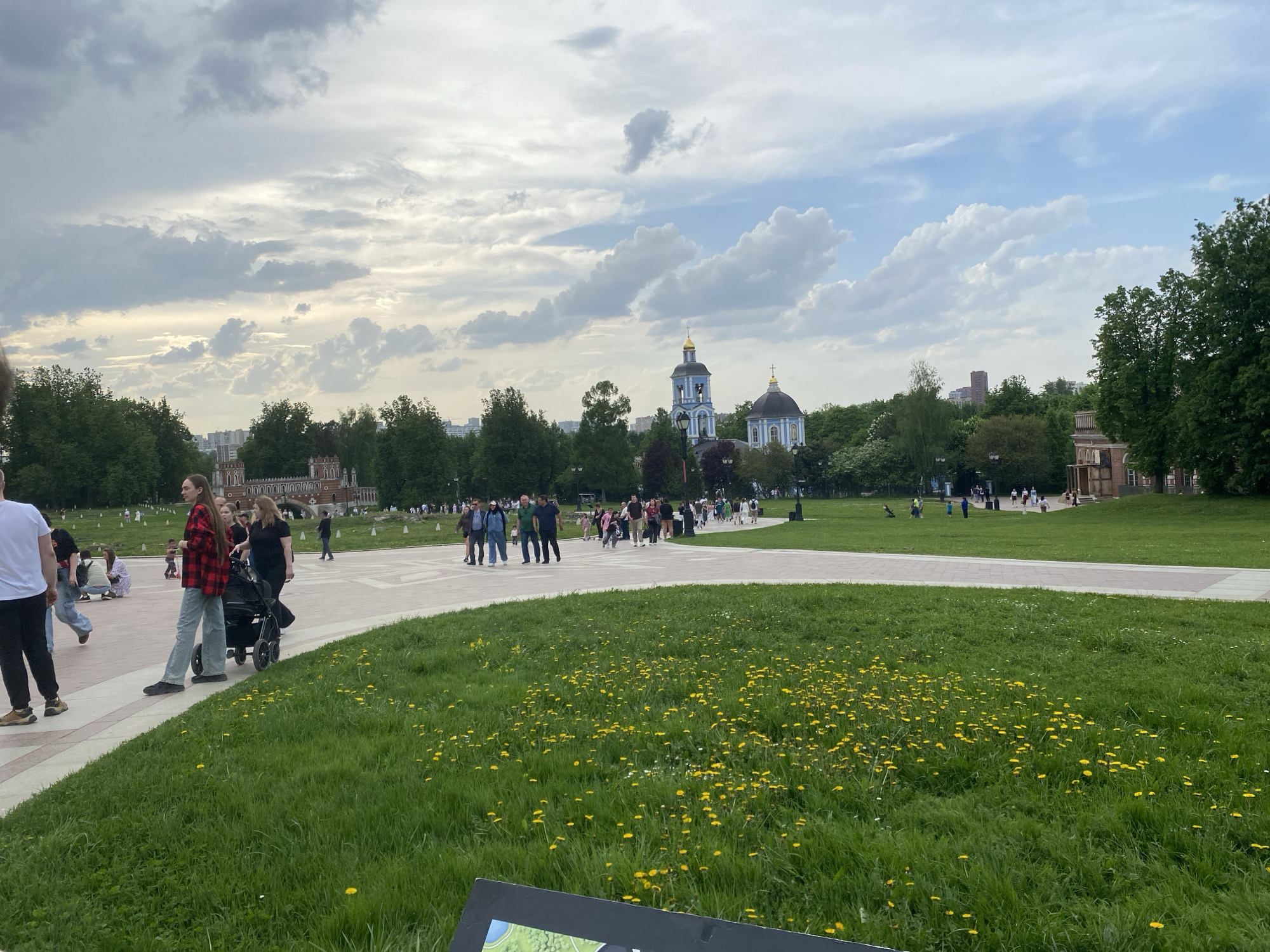 The image shows a public park, possibly in a European city, as indicated by the architecture of a church in the background. A path crosses the park with numerous people walking along it. The park consists of trees and manicured grass with a scattering of yellow flowers, most likely dandelions. In the foreground, there are people dressed in casual clothes, some with a stroller. The sky is overcast with cloudy weather conditions. The location is in the city of Unknown, Unknown.