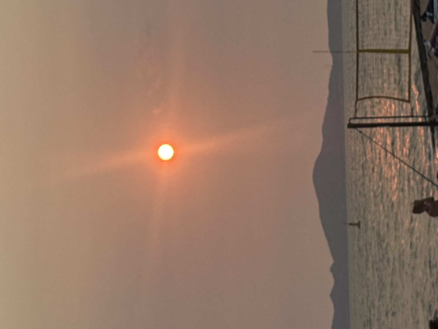 The vertically oriented image captures a coastal scene, likely during sunrise or sunset, in Pattaya City, Thailand.
The upper left portion of the frame is dominated by a bright, circular, orange-yellow sun, which emits prominent light rays that fan out into the surrounding atmosphere. The sky, which occupies the majority of the left side and top, is a hazy, reddish-orange color, suggesting diffused light conditions, possibly due to atmospheric haze or thin clouds.
On the right side of the image, a body of water, likely the sea, is visible, its surface shimmering with reflections of the warm light. A dark, undulating silhouette of a distant landmass or coastline forms a vertical separation between the sky and the water, extending downwards from the middle of the frame.
In the far right, partially visible elements of a boat or pier structure appear. These include dark, linear forms that resemble rigging, railings, or parts of a mast. A human arm and shoulder are discernible at the top right edge, indicating the presence of a person on this structure. A small, T-shaped object, possibly a buoy or navigational marker, can be seen in the water near the landmass silhouette.
The scene conveys a sense of peaceful contemplation, characteristic of a twilight hour over a tranquil marine environment. No discernible text is visible within the image.