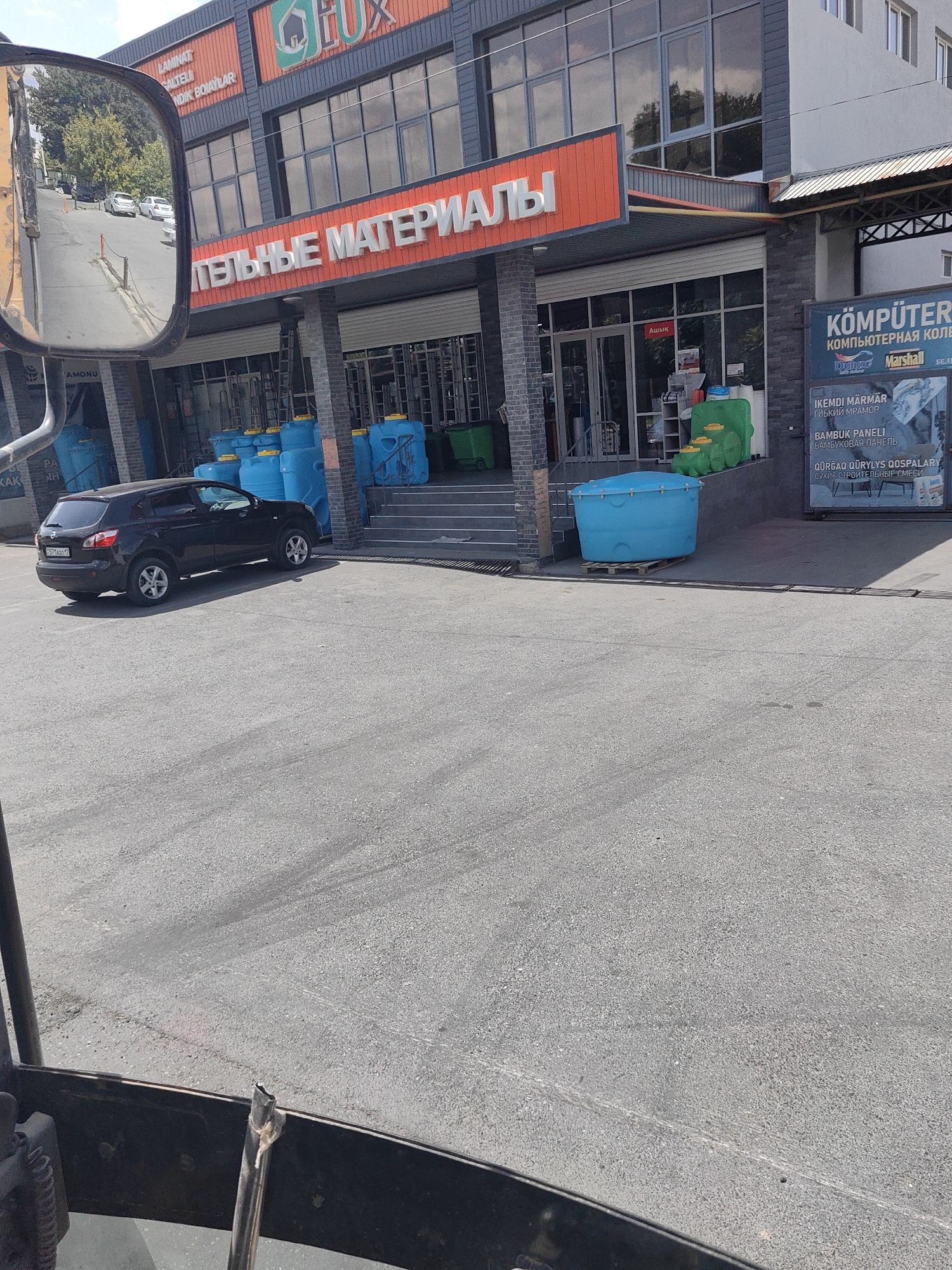 A black Nissan Qashqai is parked in front of a building with the sign "СТРОИТЕЛЬНЫЕ МАТЕРИАЛЫ" (Building Materials) in large red letters. The building appears to be a retail store selling construction supplies, with large blue water tanks and green containers visible outside. The license plate of the car is partially obscured but reads "ST116AC 17". A reflection of a street with trees and parked cars is visible in the side mirror of the vehicle. The building also has signage in Russian and Kazakh, including "КОМПЮТЕРНАЯ КОЛЛЕКЦИЯ" (Computer Collection). The scene suggests a commercial area in Shymkent, Kazakhstan, likely a street or parking lot adjacent to a business.