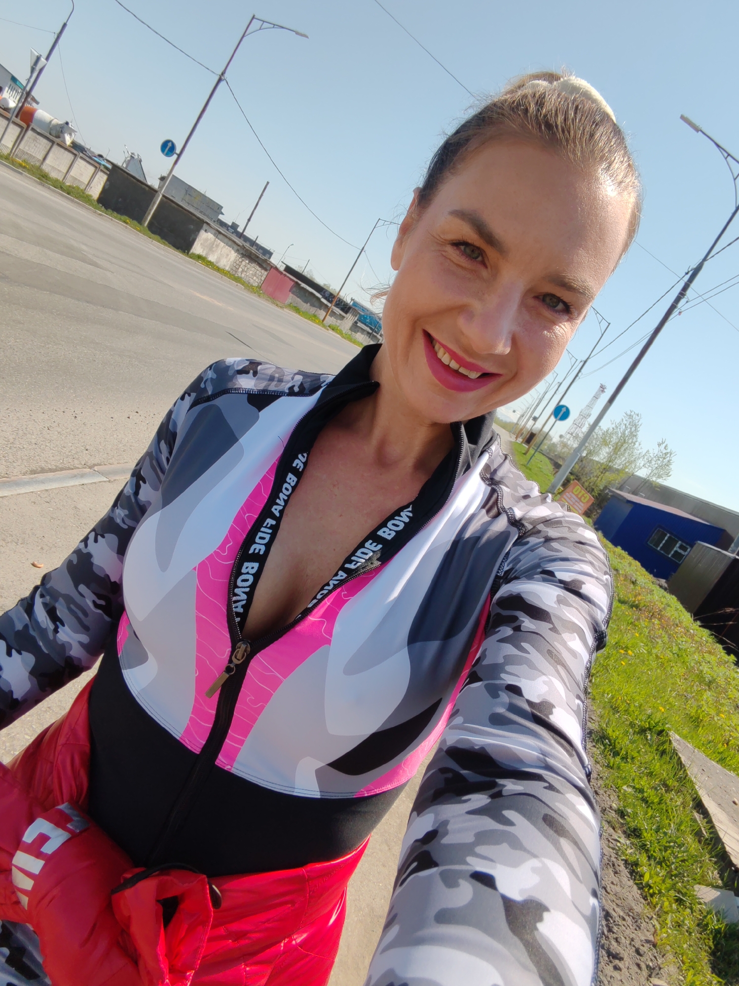 An outdoor selfie captures a fair-skinned woman smiling directly at the camera in Petropavlovsk-Kamchatsky, Russia. She has light hair pulled into a bun and wears a bright pink lipstick. Her eyes are green. She is dressed in athletic wear: a long-sleeved top featuring a camouflage pattern in gray, white, and black on the sleeves and a color-blocked torso with white, light gray, and bright pink geometric shapes over a black base. The top has a black zipper, partially undone, revealing a deeper neckline. The zipper trim features white text on a black band that reads "BONA FIDE BONA," visible in reverse on the left side of the garment (viewer's right) due to the selfie orientation. A red, possibly padded, garment with white text, possibly "BONA," is tied around her waist. Her legs are visible, covered in leggings with the same gray, white, and black camouflage pattern as her top's sleeves.
The woman stands near a paved road or asphalt surface, which dominates the lower left of the frame. The background features a clear blue sky. On the left side of the road, a concrete barrier separates the road from industrial or utility structures in the distance, including a large white cylindrical object. Overhead, multiple utility poles and power lines crisscross the sky. On the right side of the frame, a grassy embankment with scattered yellow wildflowers borders the road. Beyond the grass are several low-rise buildings, including a prominent blue structure with a white-framed window and a dark gray building with a red door. A blue circular traffic sign with a white arrow pointing right is visible above the grassy area, along with a distant lattice tower. The scene is illuminated by bright daylight.